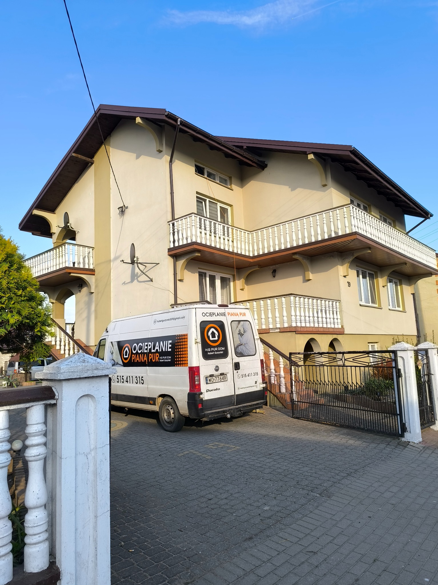 Dwupiętrowy dom z balkonami i zaparkowany biały van z logo firmy. Usługa ocieplania budynków, widok z zewnątrz na ocieplony dom i pojazd.