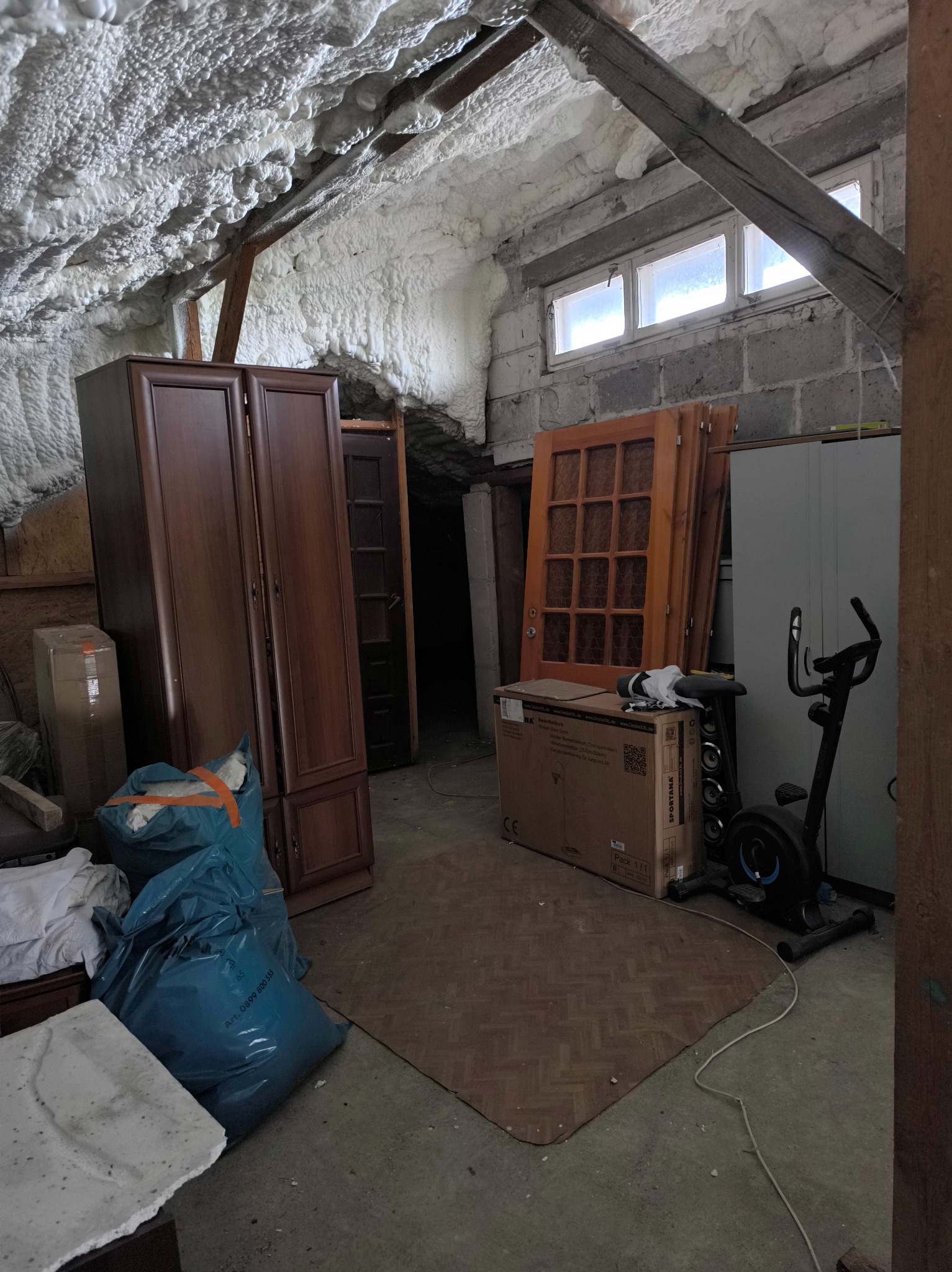 Poddasze ocieplone pianką PUR, widoczne ukośne belki stropowe i okna dachowe. Część poddasza zagracona meblami, kartonami i workami. Widoczna instalacja ocieplenia.