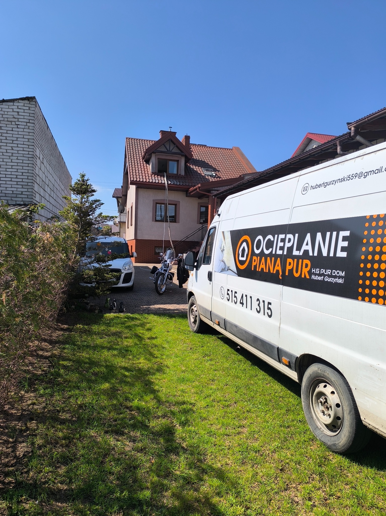 Biały van z reklamą ocieplania pianą PUR zaparkowany na trawniku przed domem. Na pojeździe widoczne dane kontaktowe i logo firmy.
