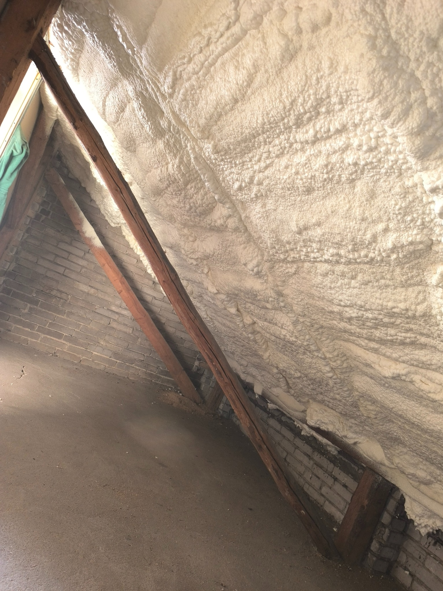 Poddasze po natrysku pianką PUR: widoczne drewniane elementy konstrukcyjne i ceglana ściana częściowo pokryta białą, gęstą pianą izolacyjną.