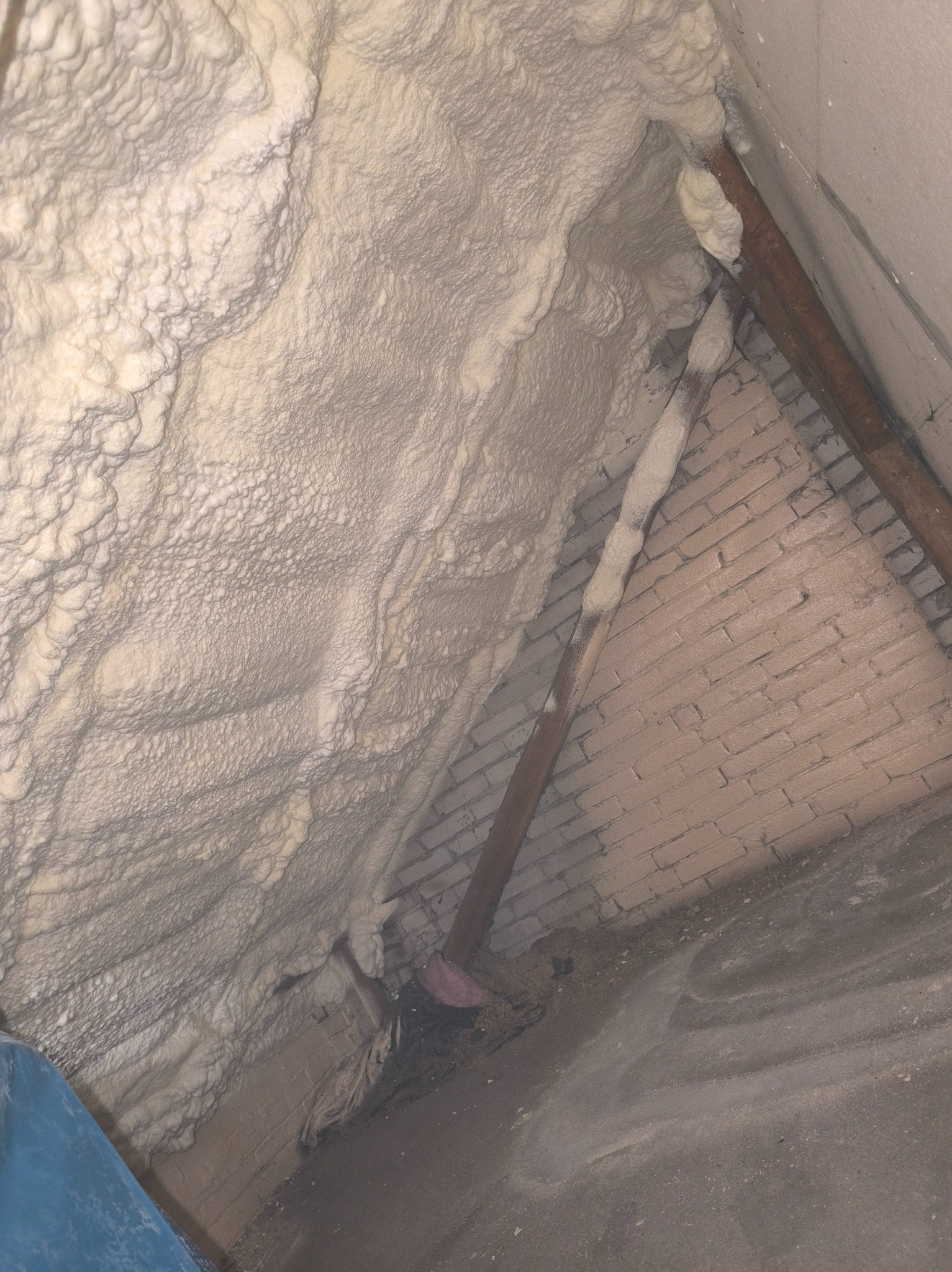 Ocieplenie poddasza pianką, widoczne cegły i drewniane belki. Aplikacja pianki zakończona, tworzy jednolitą warstwę izolacyjną.