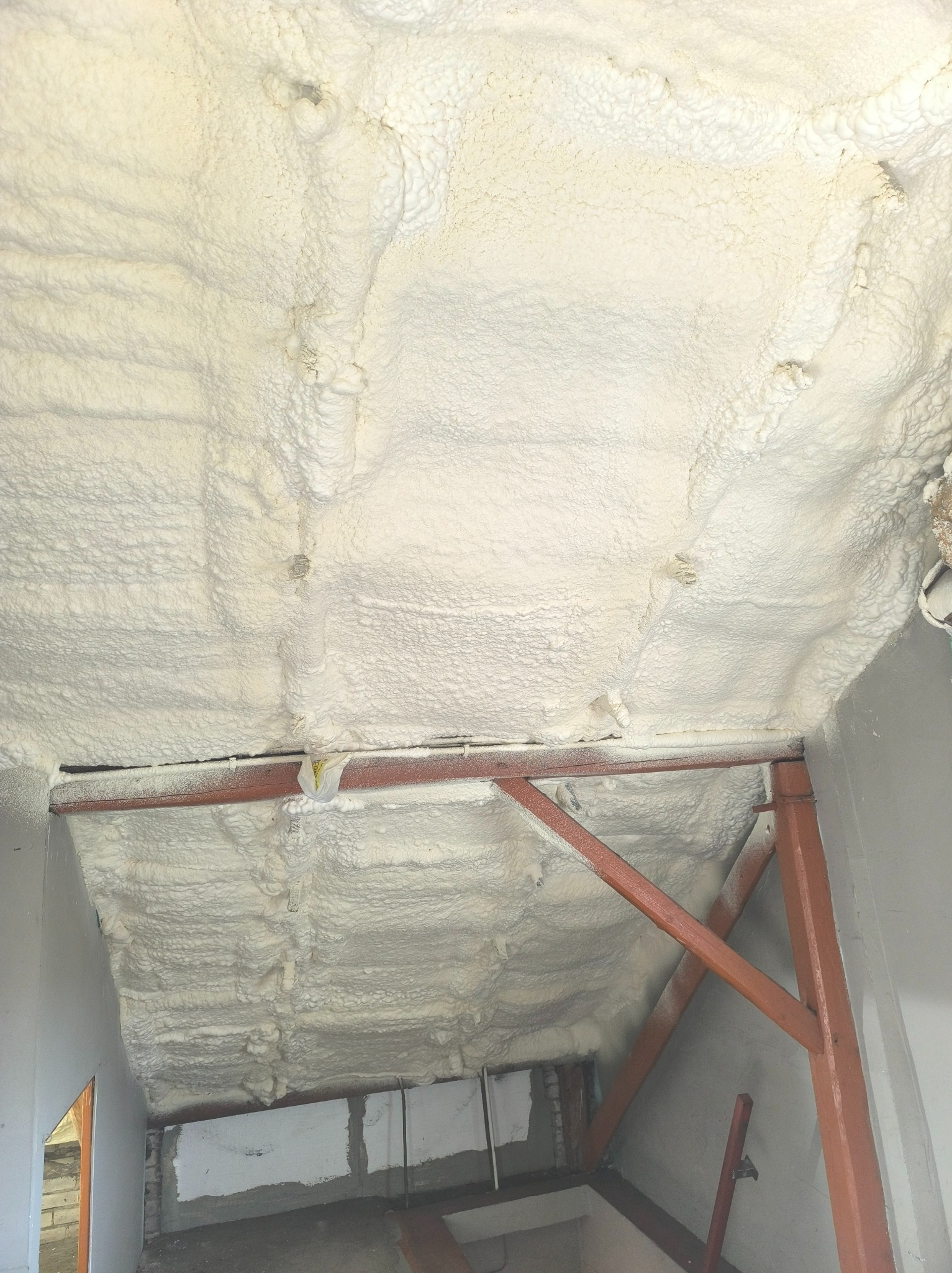 Ocieplone poddasze pianką, widoczne elementy konstrukcyjne i nierównomierna struktura izolacji, w tle fragment ściany i otworu.