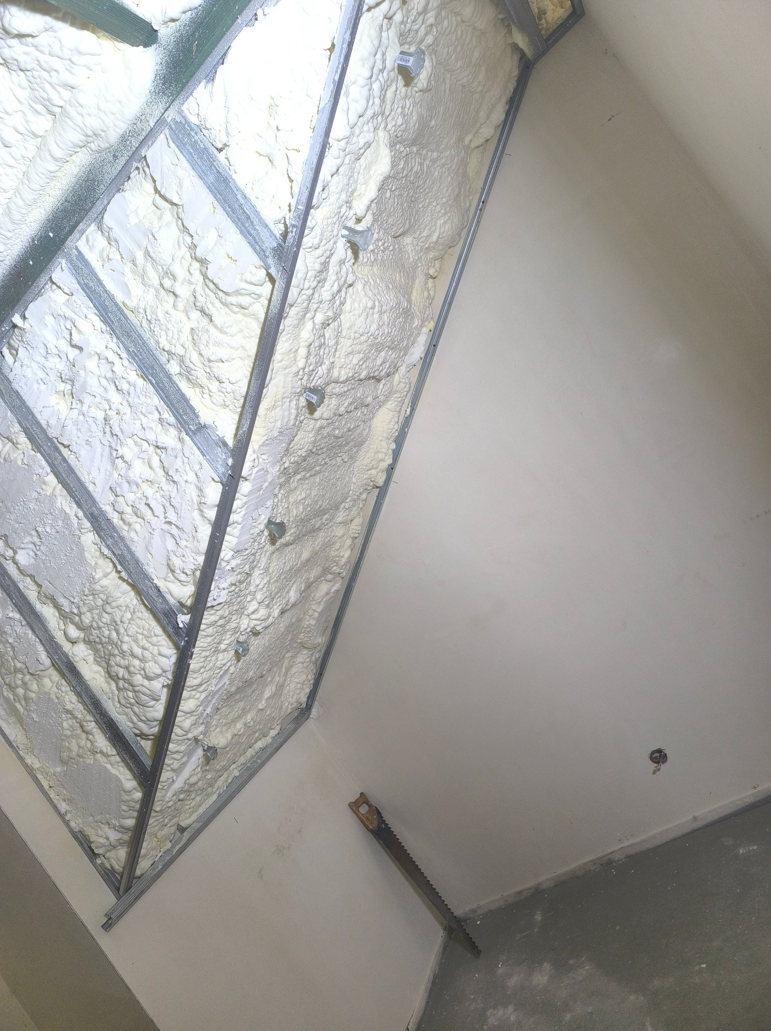 Ocieplenie poddasza pianką w metalowej konstrukcji, widoczna piła do cięcia. Biała piana wypełnia przestrzeń między profilami. Ściana w tle pomalowana na biało.