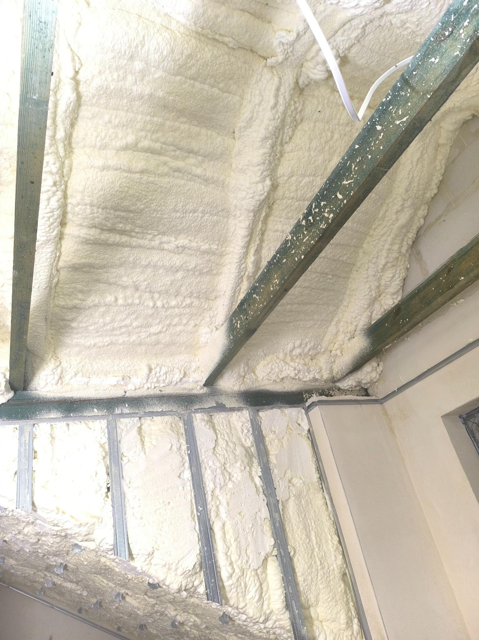 Ocieplenie poddasza pianką poliuretanową, widoczne metalowe profile i drewniane belki. Biała, gęsta struktura piany wypełnia przestrzeń.