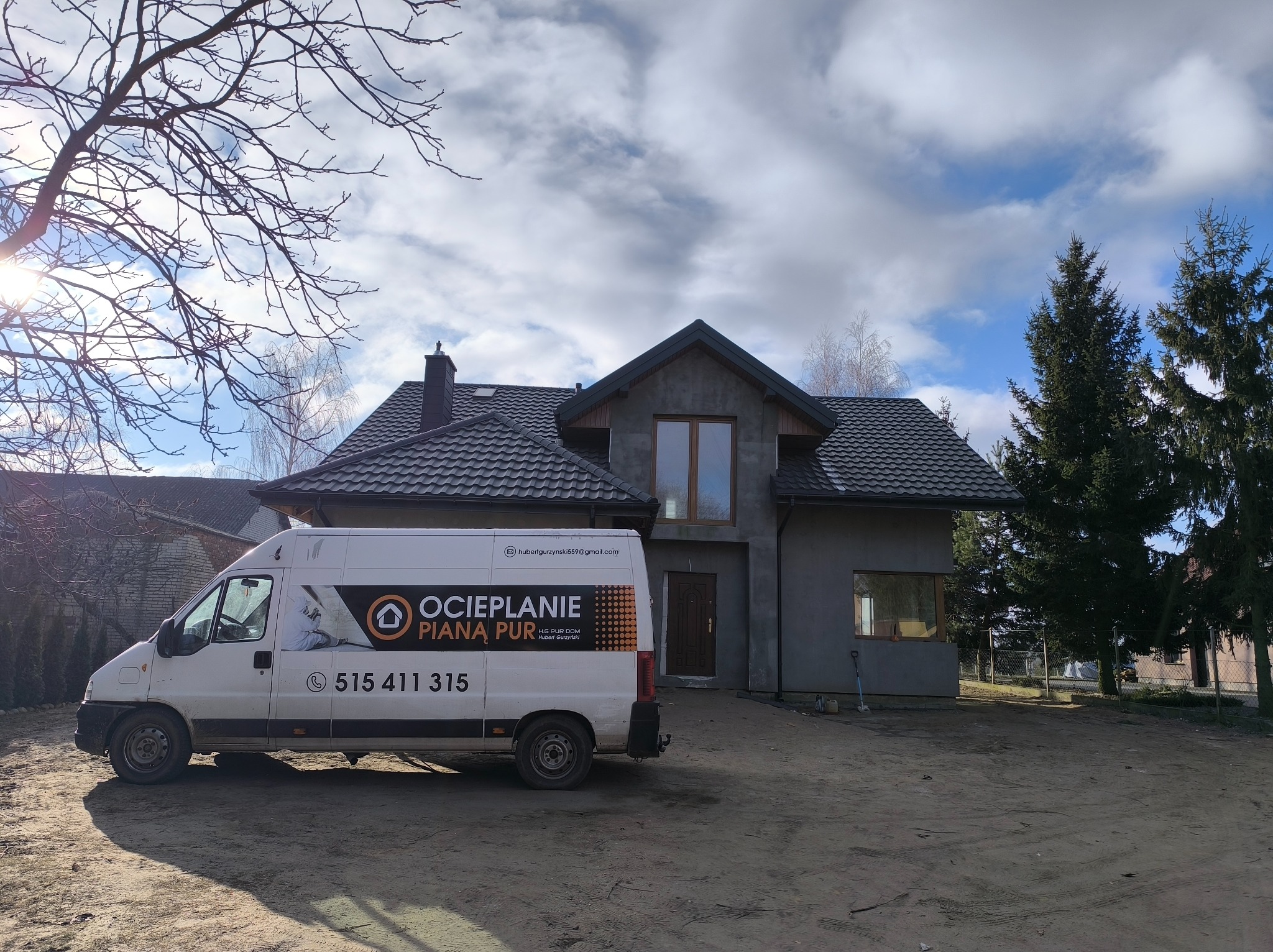 Ocieplony dom jednorodzinny z szarą elewacją, widoczny samochód dostawczy z reklamą ocieplania pianą PUR. Słoneczny dzień, częściowo zachmurzone niebo.