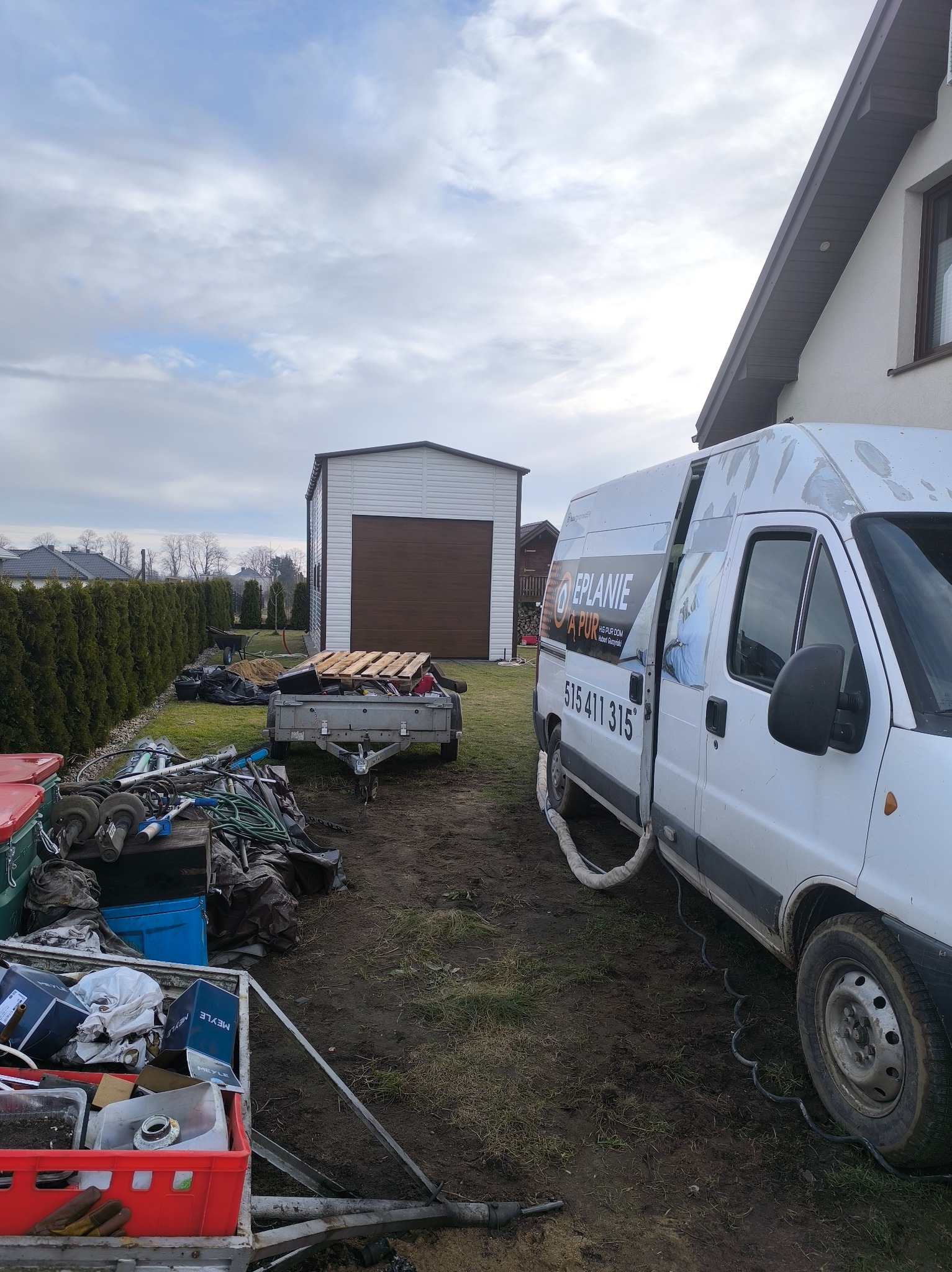 Mobilny punkt aplikacji pianki PUR z widocznym wężem i przyczepką na posesji. W tle garaż blaszany i budynek mieszkalny.