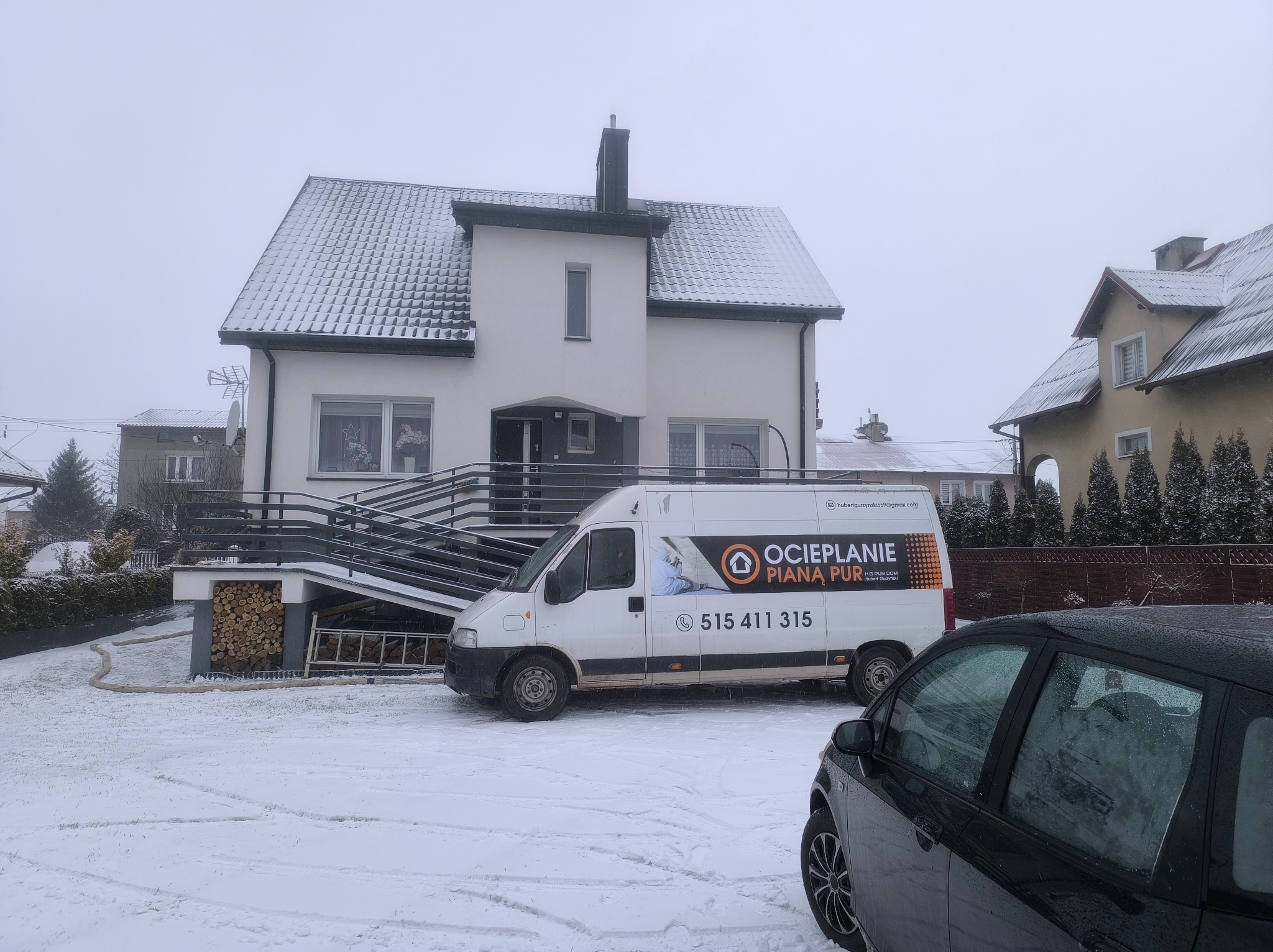 Ośnieżony dom jednorodzinny z zaparkowanym samochodem firmy ociepleniowej. Zimowa sceneria, szara pogoda. Widoczny stos drewna i drabina.