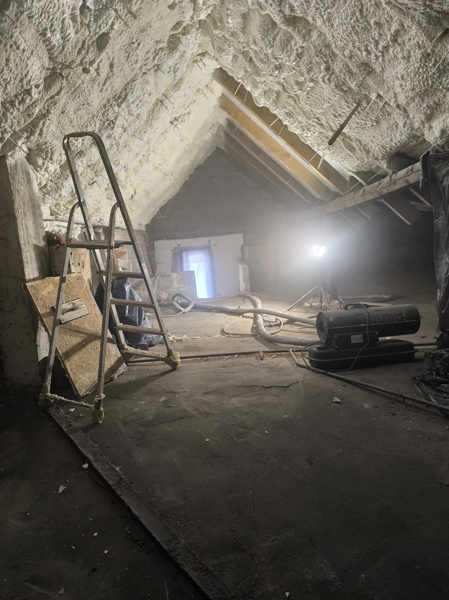 Poddasze po ociepleniu pianką PUR: widoczna struktura pianki na skośnych ścianach, drabina oparta o ścianę, sprzęt do natrysku i oświetlenie budowlane.