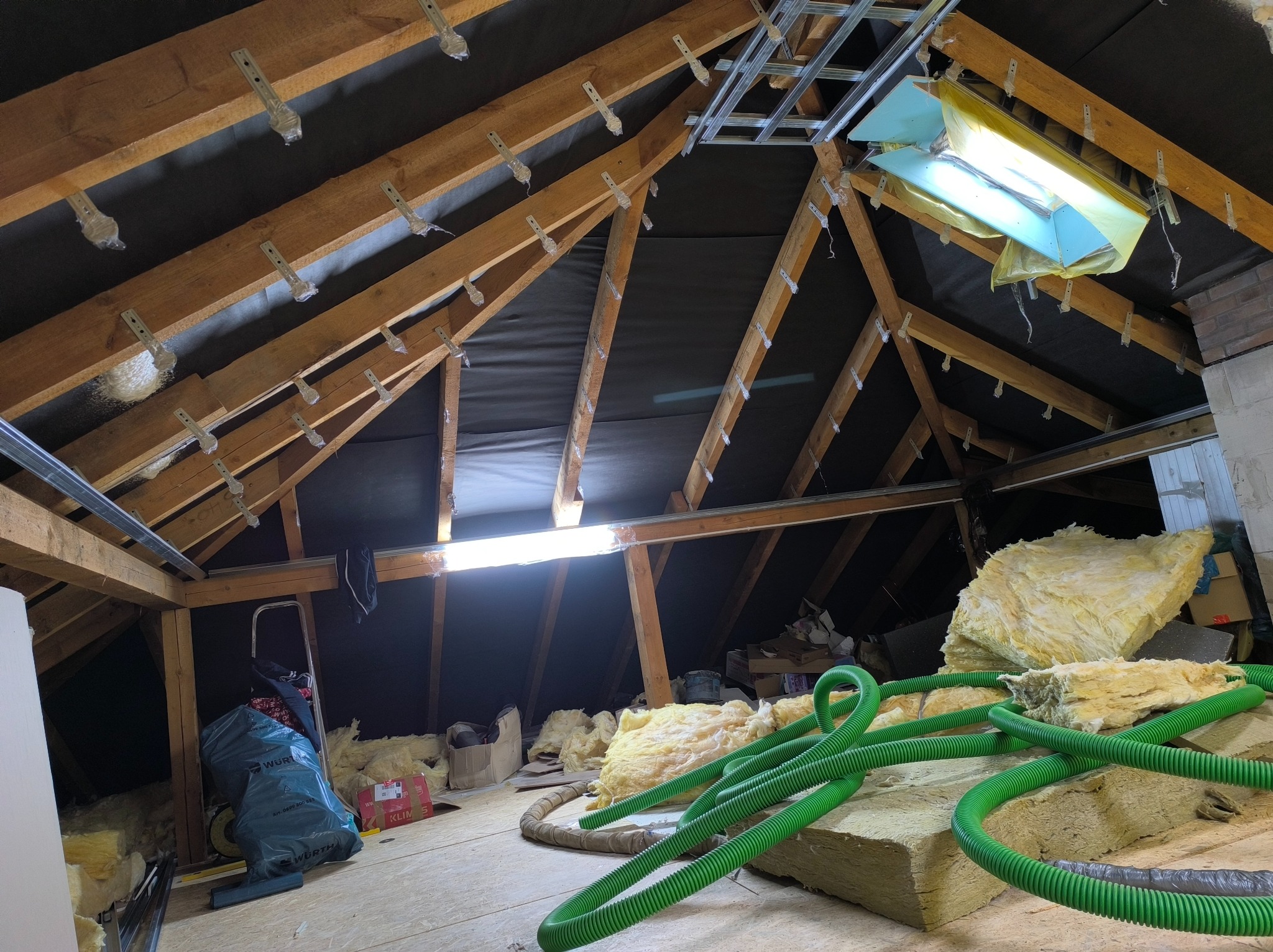 Poddasze w trakcie prac: widoczne krokwie z membraną, oświetlenie robocze, materiały izolacyjne (wełna mineralna) i przewody wentylacyjne. Etap przygotowania do ocieplenia.
