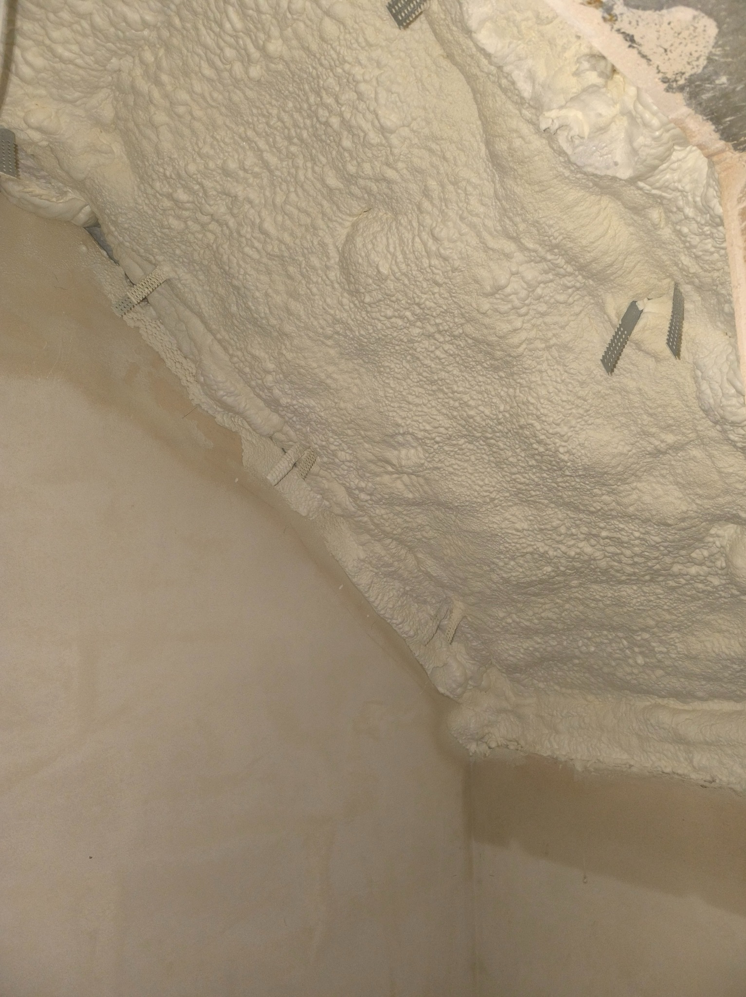 Ściana i strop po natrysku pianki izolacyjnej. Widoczne metalowe elementy mocujące. Szorstka faktura piany. Izolacja poddasza.