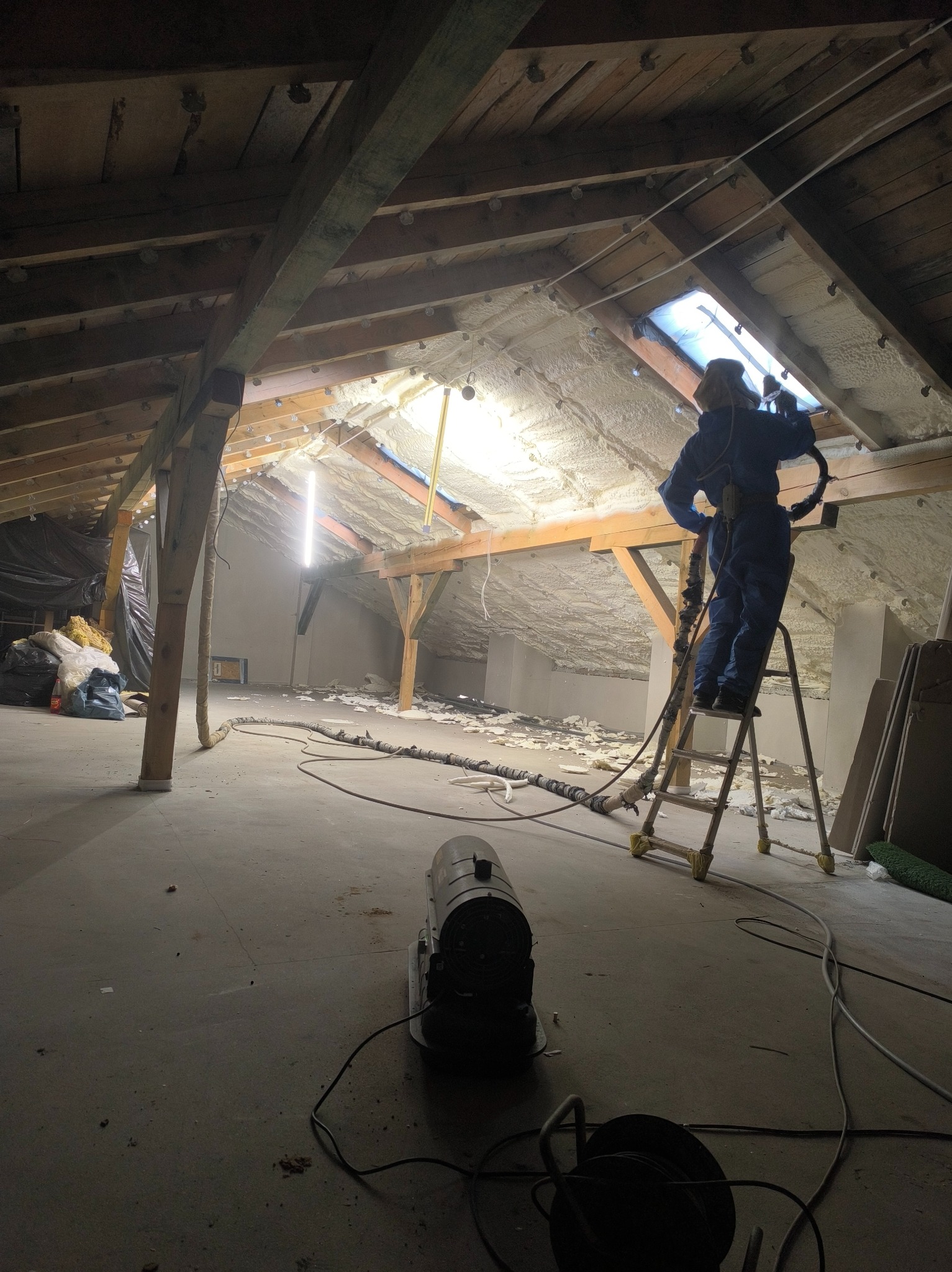 Pracownik w kombinezonie na drabinie aplikuje izolację natryskową na poddasze. Widoczne drewniane belki, oświetlenie i sprzęt do natrysku.