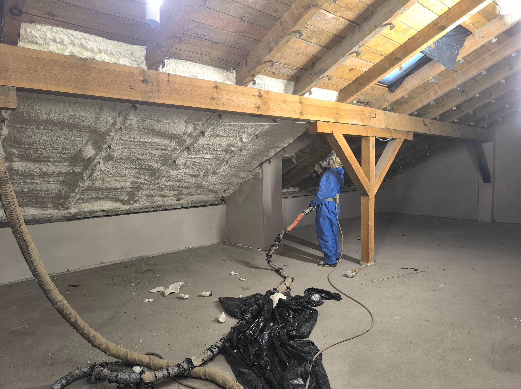 Pracownik w kombinezonie aplikuje pianę izolacyjną na poddaszu. Widoczne drewniane elementy konstrukcyjne i gruba warstwa piany. Częściowo ocieplony strop.