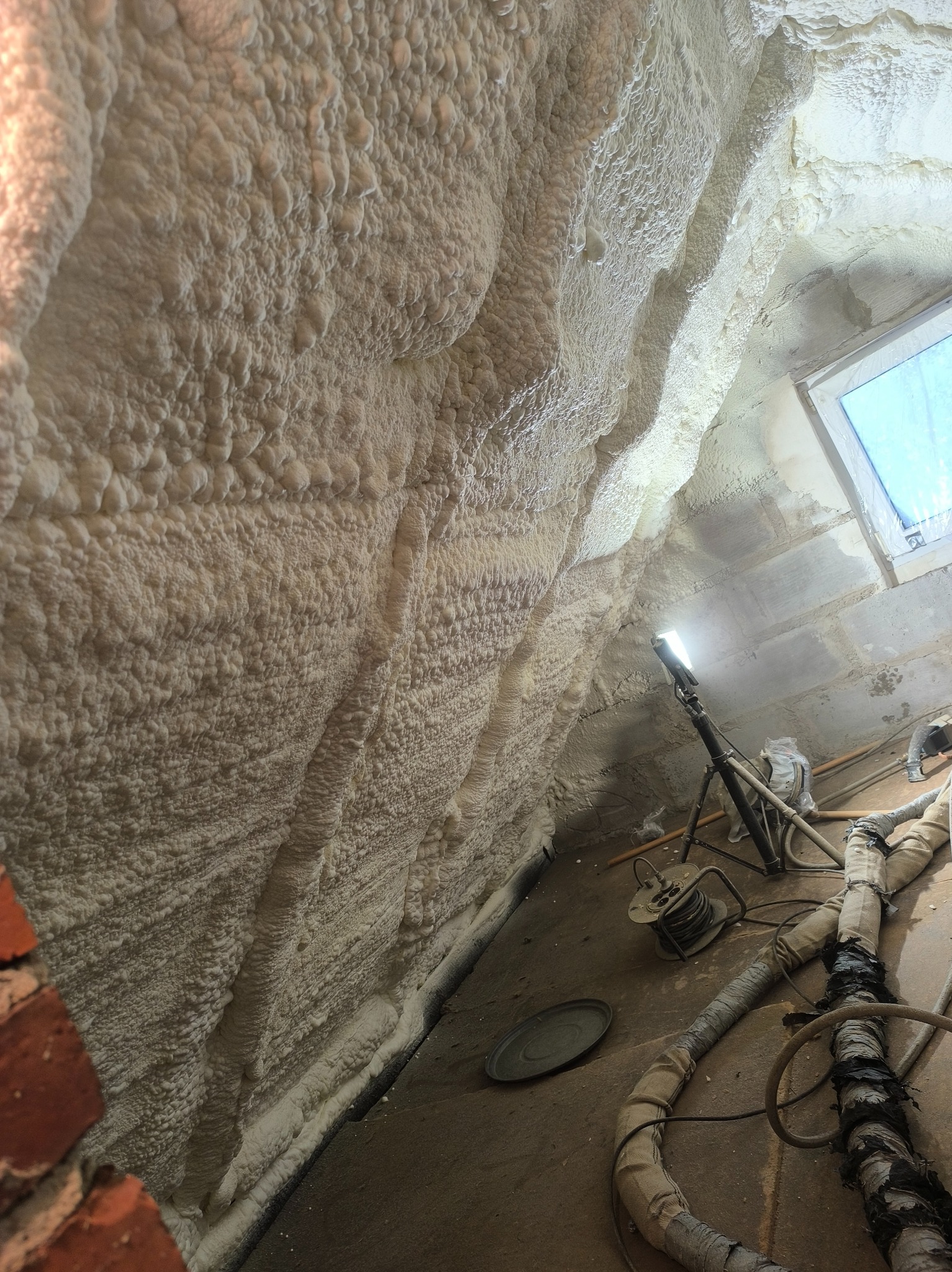 Ściana i sufit po natrysku pianki izolacyjnej, widoczne narzędzia i lampa oświetlająca. Detal faktury piany, okno w tle. Wykończona izolacja poddasza.