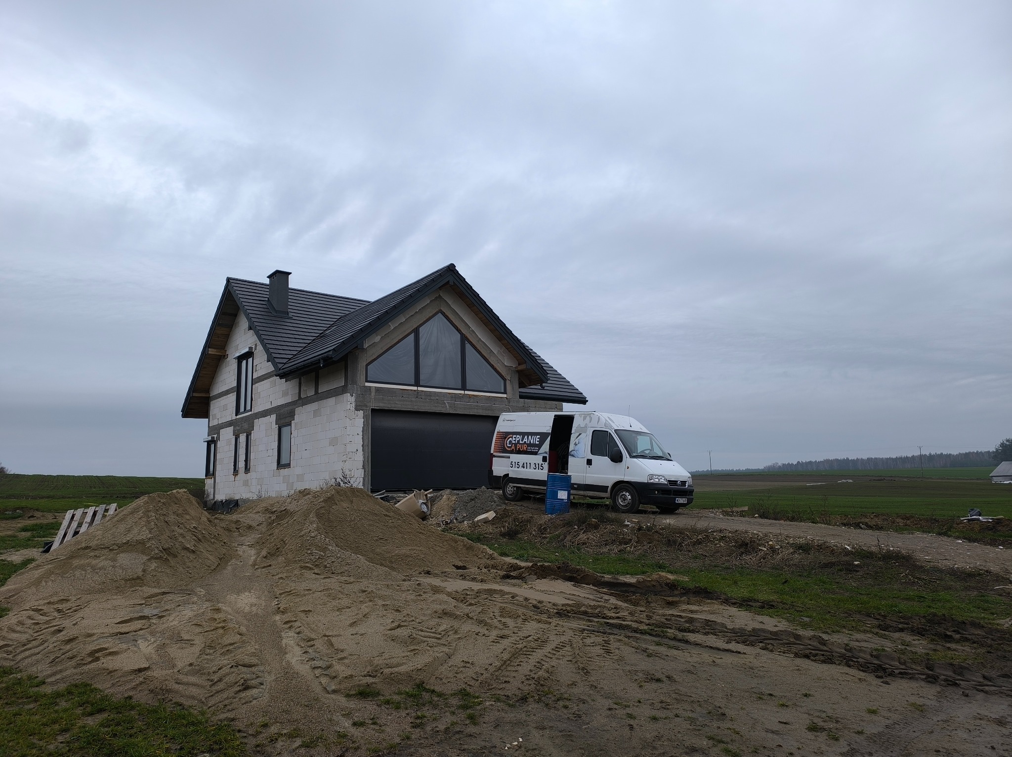 Budowa domu z pustaków, widoczny samochód firmy ociepleniowej z otwartymi drzwiami. Na pierwszym planie hałda piasku. Pochmurne niebo.