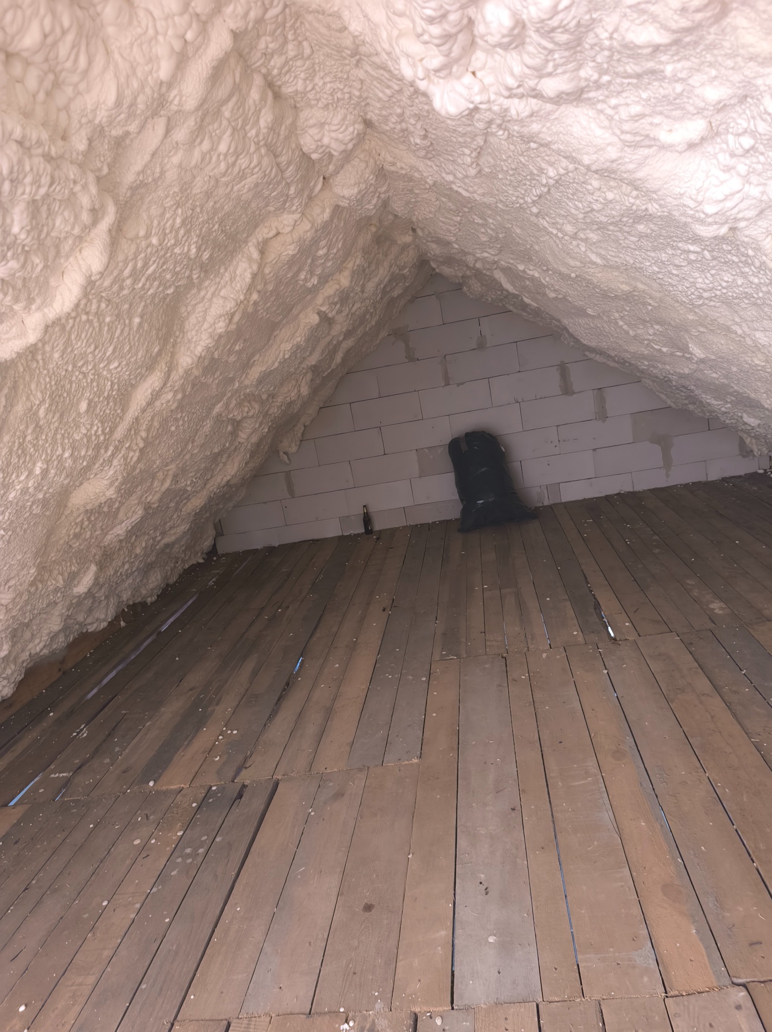 Strych z drewnianą podłogą i ociepleniem pianką na skośnych ścianach oraz suficie. Widoczny worek i butelka przy ścianie z bloczków.