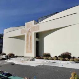 Biały budynek z nowoczesną elewacją, zdobiony malowidłem religijnym, zabezpieczony folią malarską podczas prac wykończeniowych.