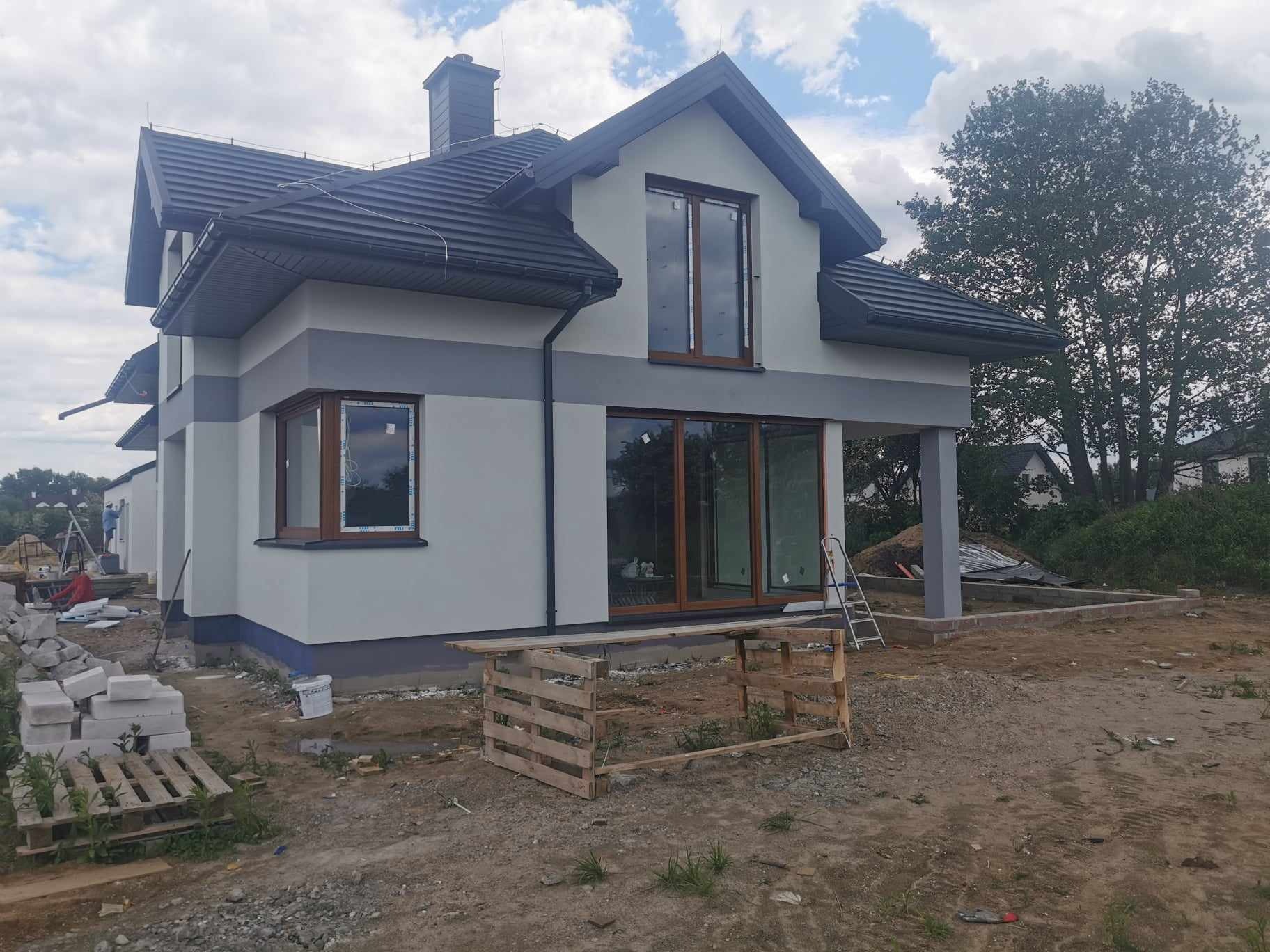 Dwukondygnacyjny dom z szarą elewacją, ciemną dachówką i brązowymi oknami, widoczny w trakcie prac wykończeniowych, z paletami i materiałami budowlanymi na pierwszym planie.