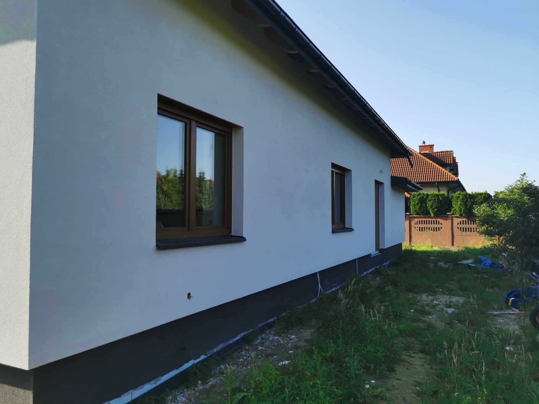 Ocieplony dom jednorodzinny z jasną elewacją, brązowymi oknami i ciemną podmurówką, widok z boku na tle zieleni i sąsiedniego domu z czerwoną dachówką.