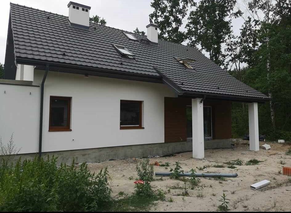 Nowoczesny dom jednorodzinny z białą elewacją, brązowymi oknami i ciemnoszarą dachówką, widok z boku na ogród w trakcie prac wykończeniowych.