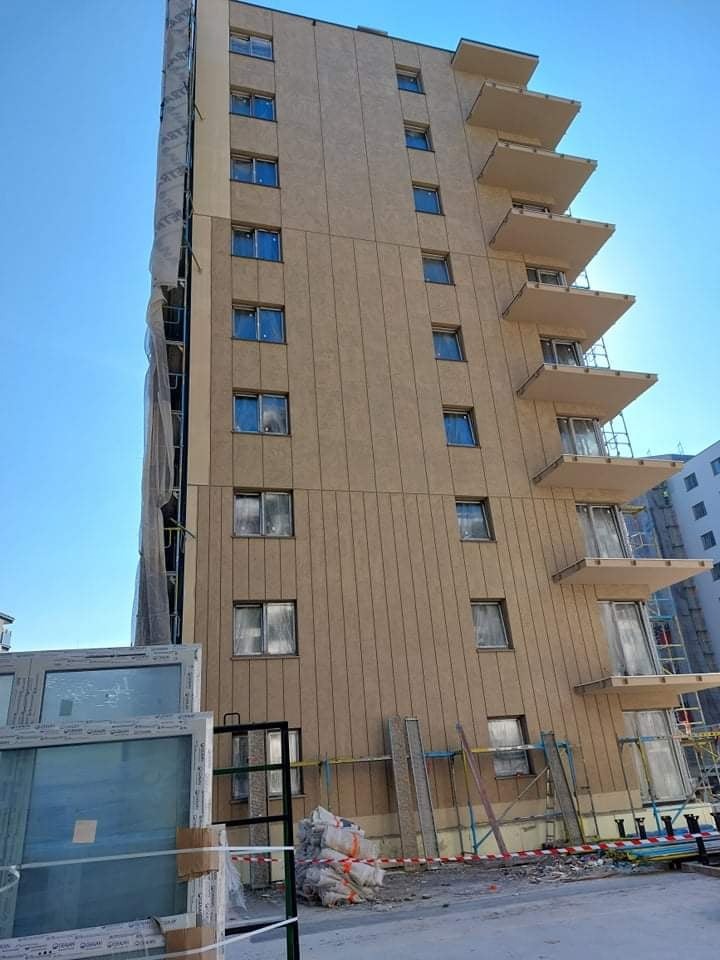 Wielopiętrowy budynek w trakcie prac elewacyjnych, widoczne panele elewacyjne, balkony i okna, z jednej strony budynku siatka ochronna, na dole składowane okna i materiały budowlane.