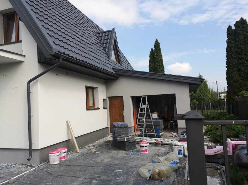 Pomalowana na szaro elewacja domu jednorodzinnego z ciemną dachówką, widoczne przygotowania do dalszych prac: folia ochronna, drabina, farby i styropian na podłożu.