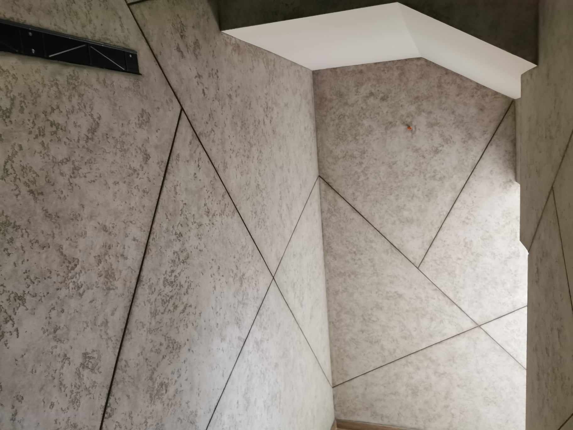 Wnętrze z wykończoną ścianą o fakturze betonu, podzieloną czarnymi listwami na geometryczne panele, widoczna kratka wentylacyjna i fragment sufitu.