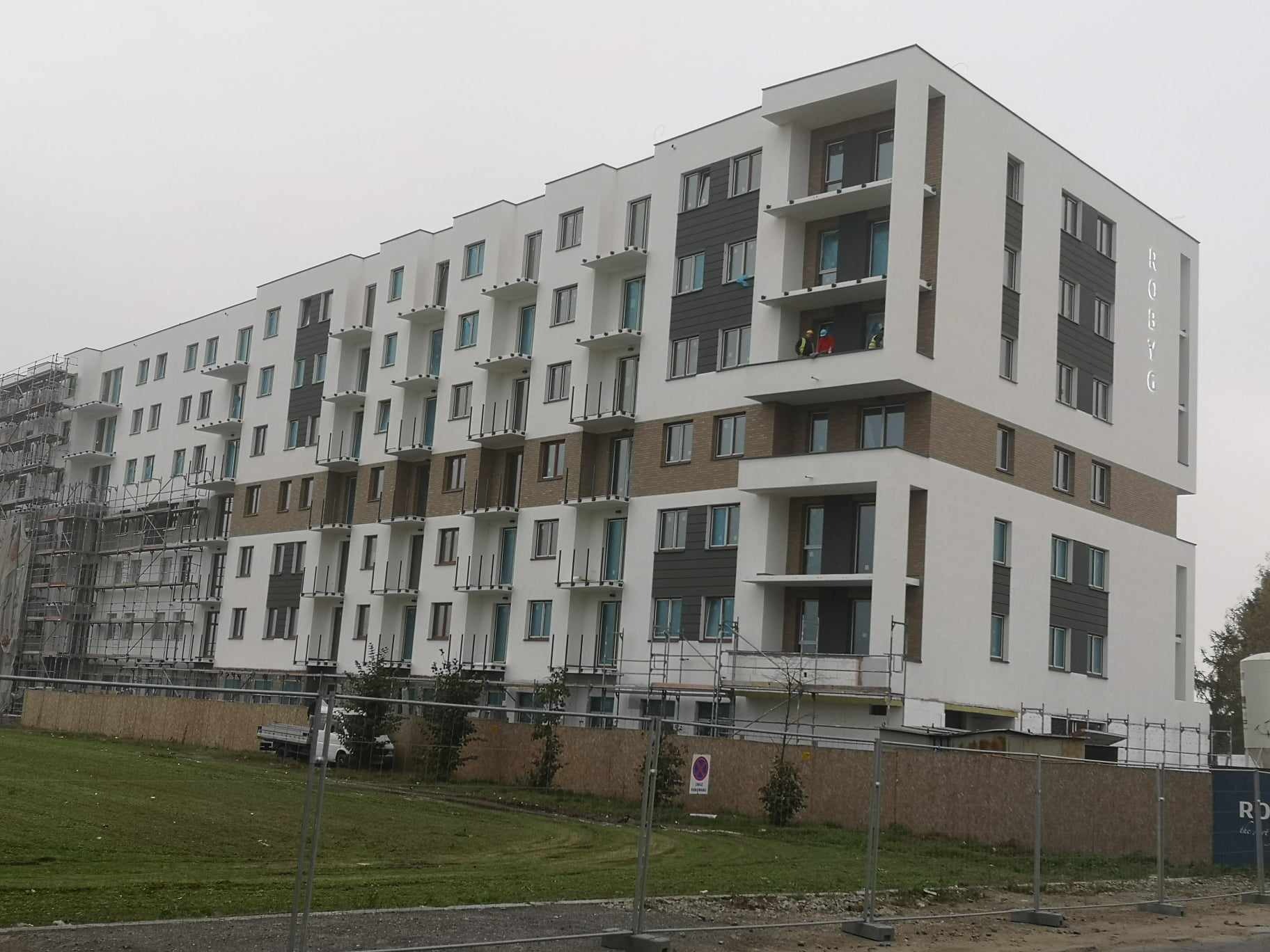 Nowoczesny budynek wielorodzinny z białą elewacją, szarymi panelami i balkonami, częściowo w rusztowaniach, z dwoma pracownikami na balkonie w trakcie prac.