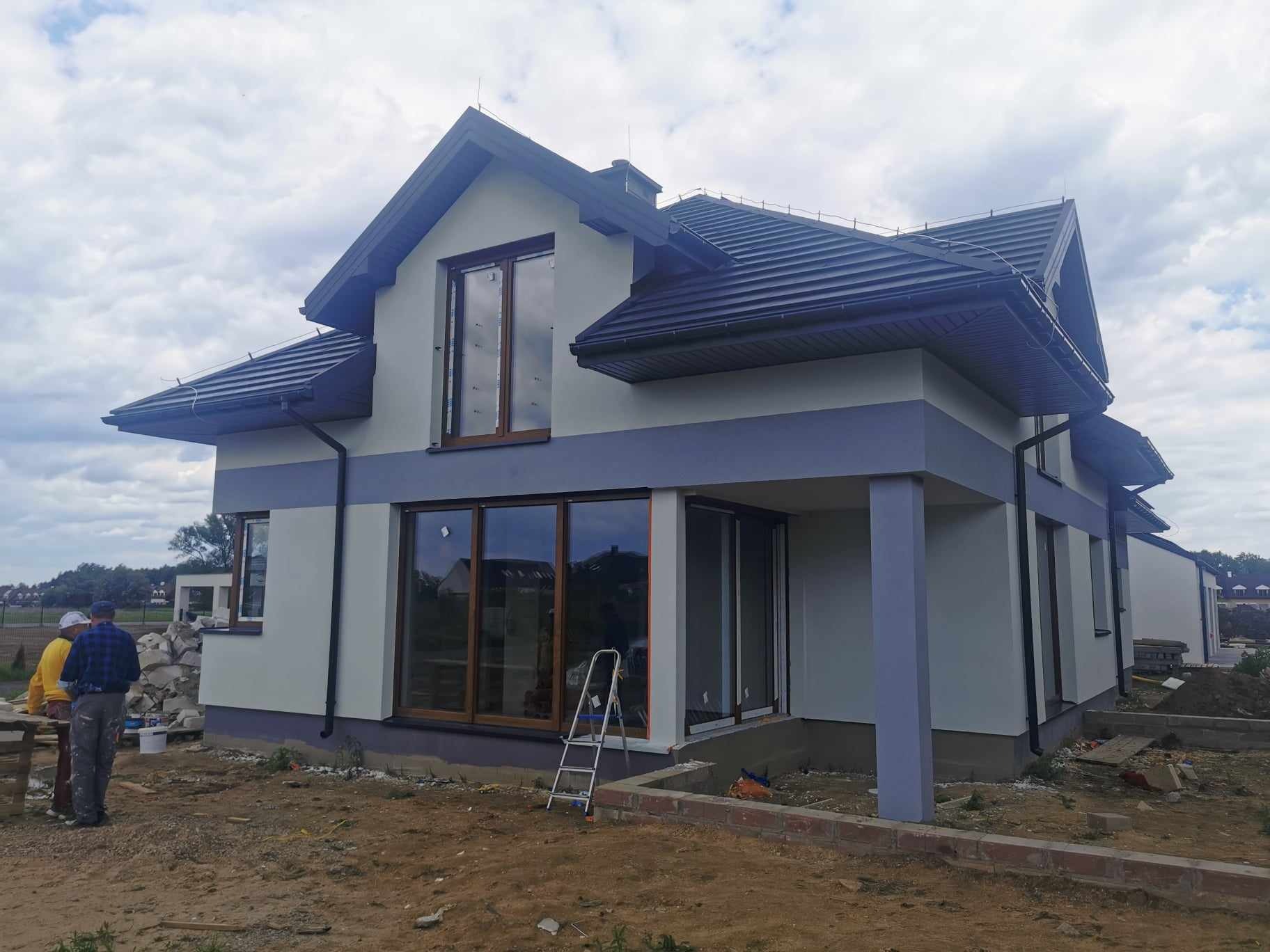 Dwukondygnacyjny dom z szarą elewacją i ciemną dachówką, widoczne okna z brązowymi ramami i metalowa drabina oparta o ścianę, w tle dwóch pracowników budowlanych.