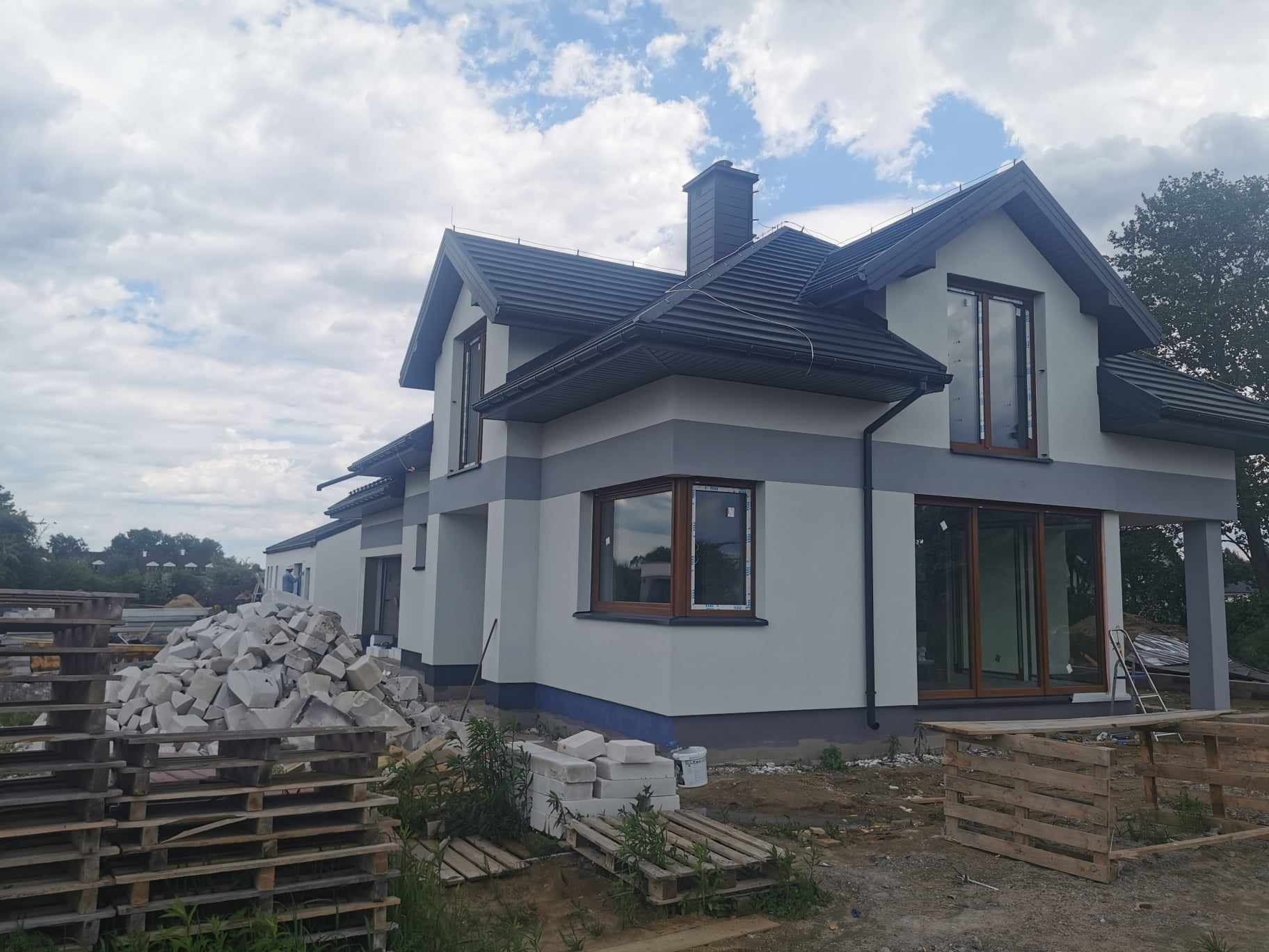 Nowoczesny dom jednorodzinny z szarą elewacją w trakcie wykończenia, widoczne palety z materiałami budowlanymi i stos białych bloczków na trawniku.