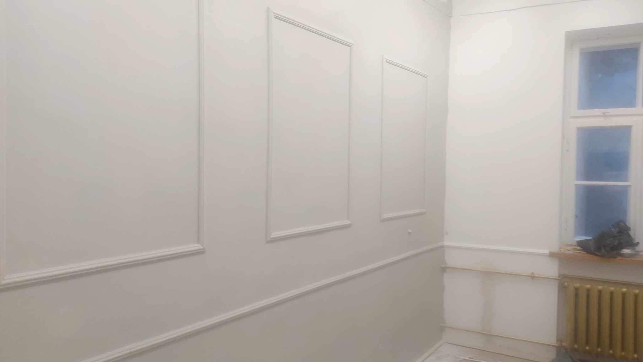 Białe ściany z dekoracyjnymi listwami i panelami ściennymi, grzejnik pod oknem, wnętrze po malowaniu.