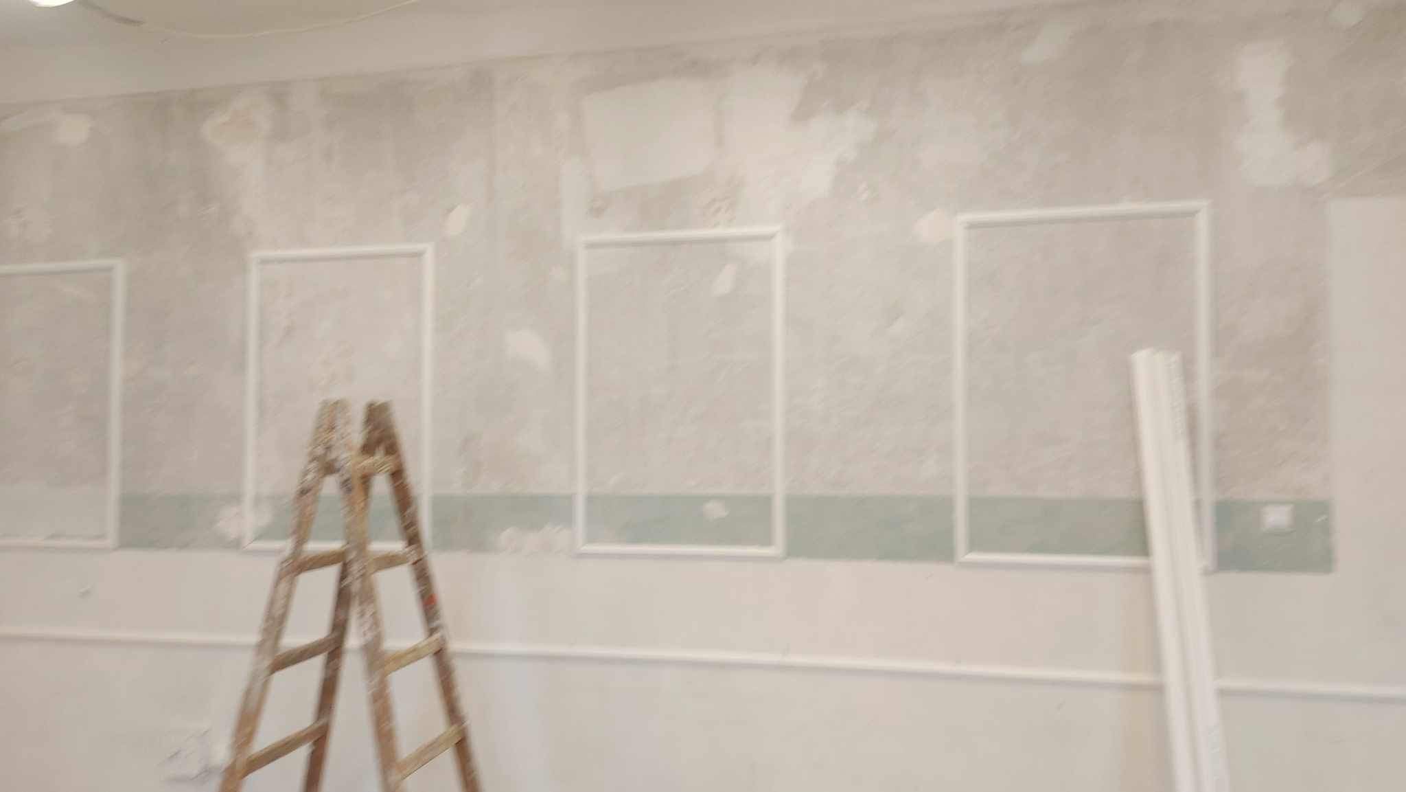 Ściana w trakcie dekorowania z zamontowanymi białymi listwami tworzącymi ramy, widoczna drewniana drabina i listwy przypodłogowe