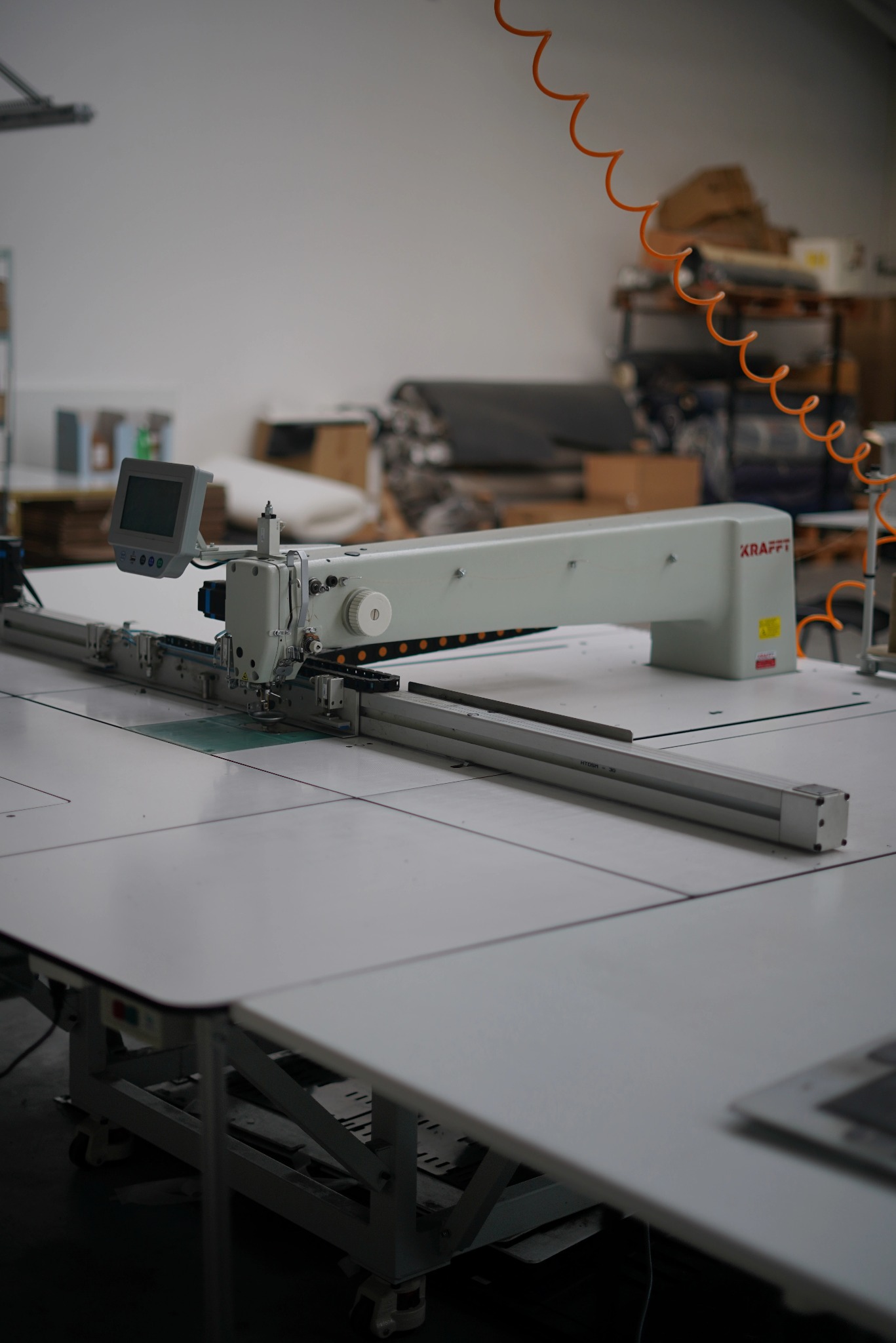 Profesjonalna maszyna krojcza marki KRAFFT z panelem sterowania, umieszczona na białym stole w szwalni, w tle widoczne materiały i regały.