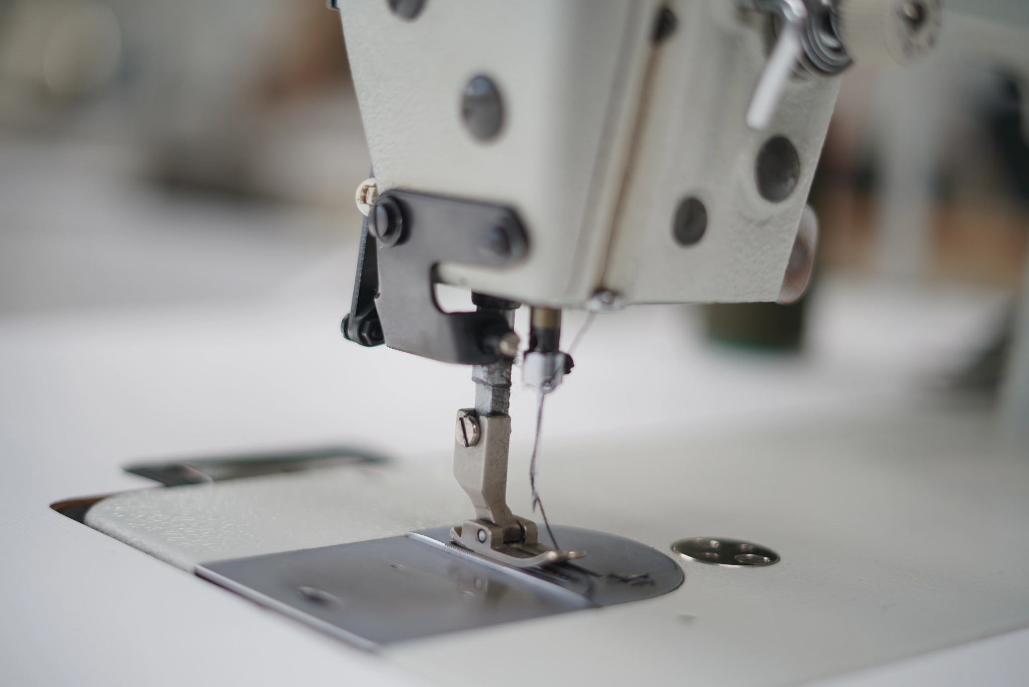 Zbliżenie na stopkę i igłę przemysłowej maszyny do szycia na tle białego blatu, z widocznym fragmentem mechanizmu i niewyraźnym tłem szwalni.