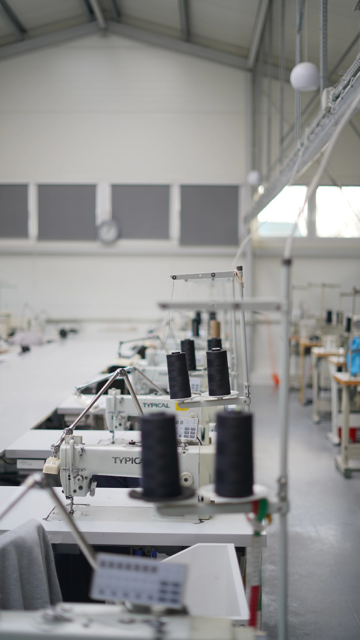 Rząd białych maszyn do szycia marki Typical z czarnymi szpulami nici w jasnej, przestronnej szwalni z oknami i szarymi panelami na ścianach.