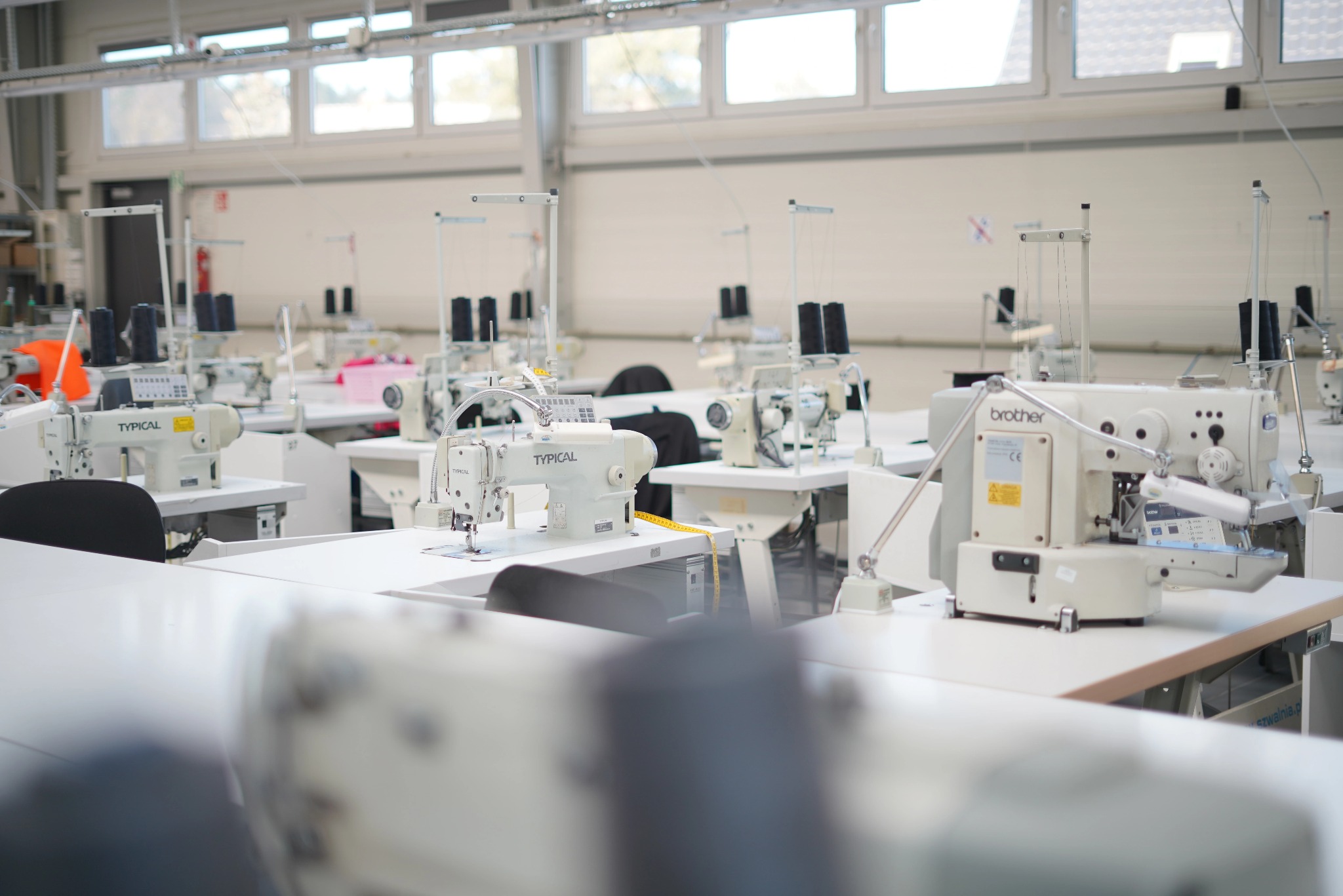 Jasne wnętrze szwalni z rzędami białych stołów i maszynami do szycia, w tle okna wpuszczające naturalne światło.