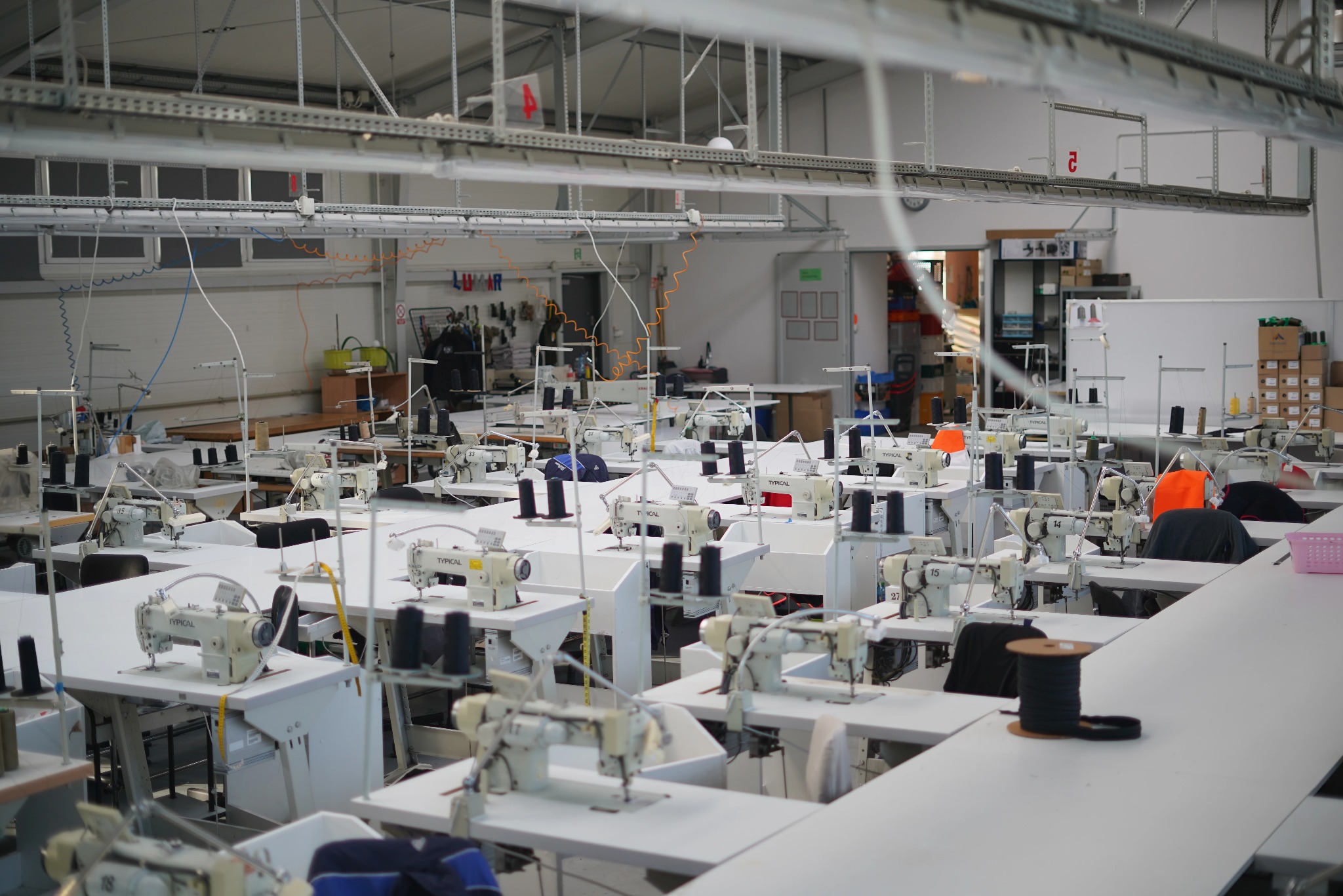 Przestronna szwalnia z rzędami białych stołów i maszyn do szycia Typical, czarne szpule nici na stołach, widoczne wieszaki z ubraniami w tle.
