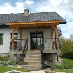 Tomek - Dom jednorodzinny z zadaszonym tarasem, widoczne schody wejściowe z kamienia, czarna balustrada i drewniana konstrukcja zadaszenia. Na dachu szara dachówka ceramiczna, a w tle błękitne niebo...