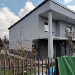 Tomek - Nowoczesny, szary dom z balkonem i elewacją wykończoną kamieniem, widziany zza metalowego ogrodzenia z zielonym materiałem i niebieską folią w tle.