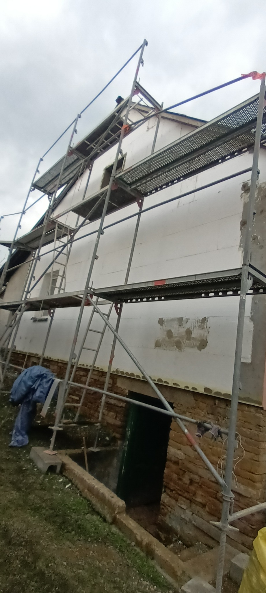 Dom w trakcie ocieplania styropianem, widoczne rusztowania i odsłonięta ceglana podmurówka, częściowo zakryta folią budowlaną.