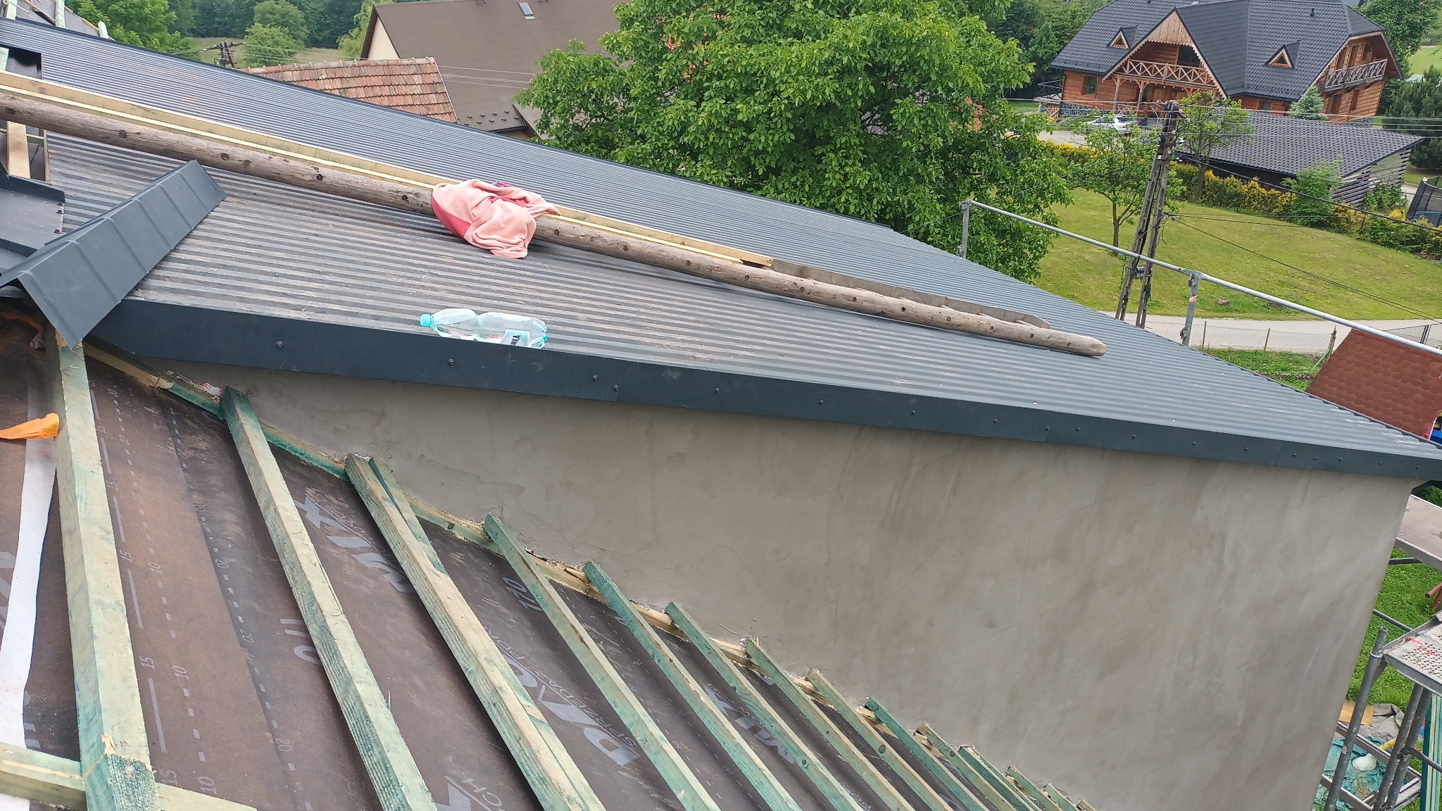 Montaż dachu z blachy trapezowej w trakcie realizacji, widoczne drewniane łaty, membrana dachowa, butelka wody i ręcznik na dachu, w tle zabudowa jednorodzinna.