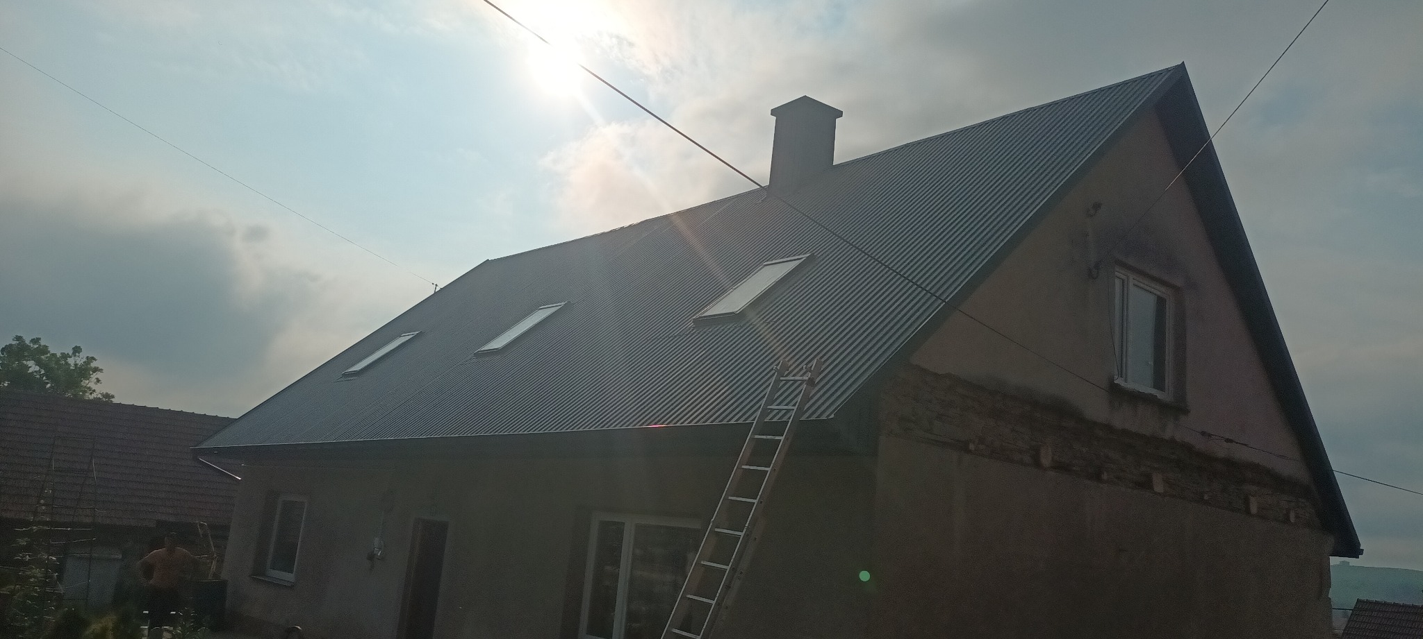 Dom z nowym, szarym dachem z blachy trapezowej, widoczne trzy okna dachowe i komin, drabina oparta o ścianę, fragment elewacji w trakcie prac wykończeniowych.