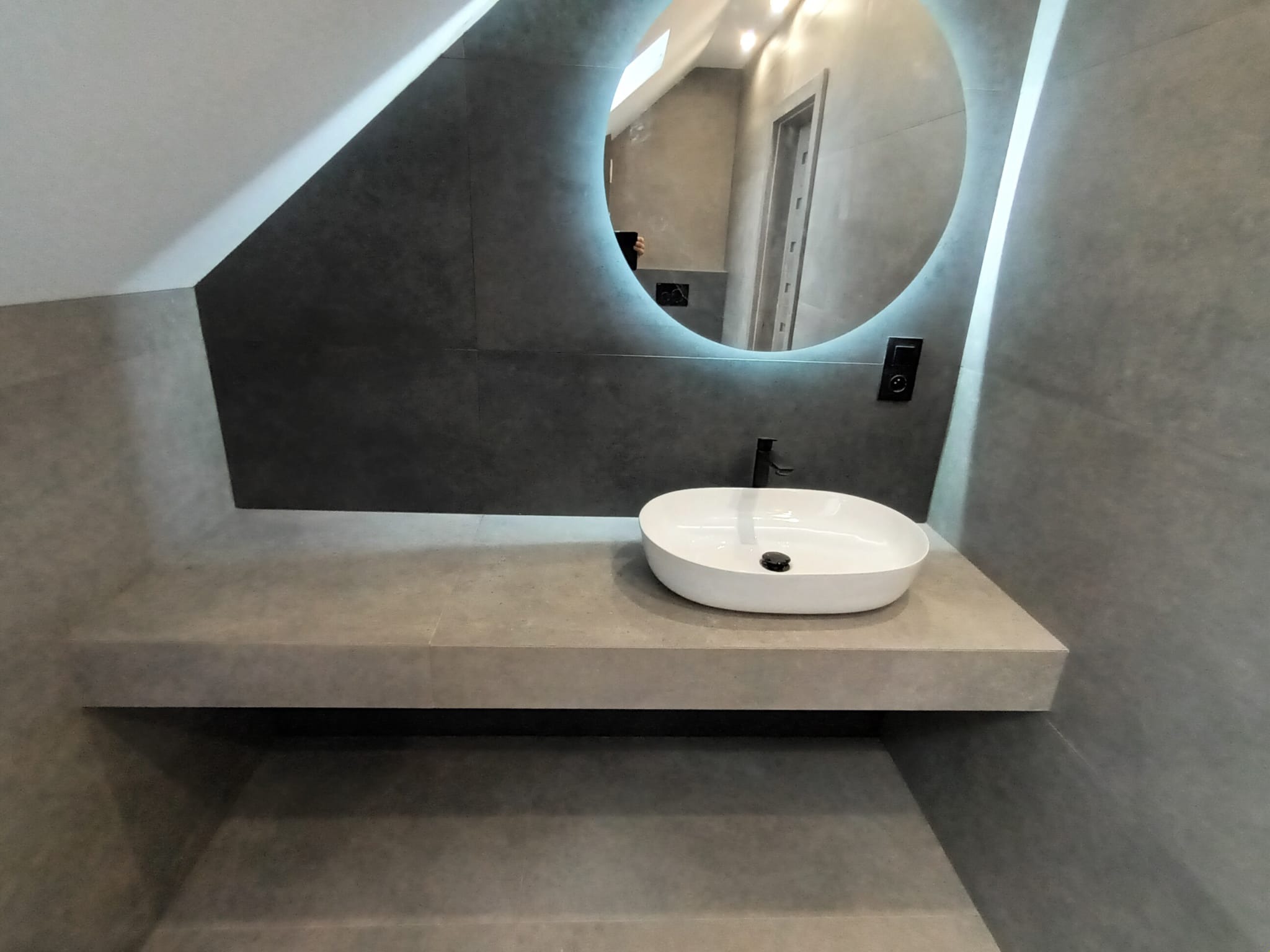 Nowoczesna łazienka na poddaszu z szarą, kamienną umywalką nablatową, okrągłym lustrem z podświetleniem LED i czarną armaturą.