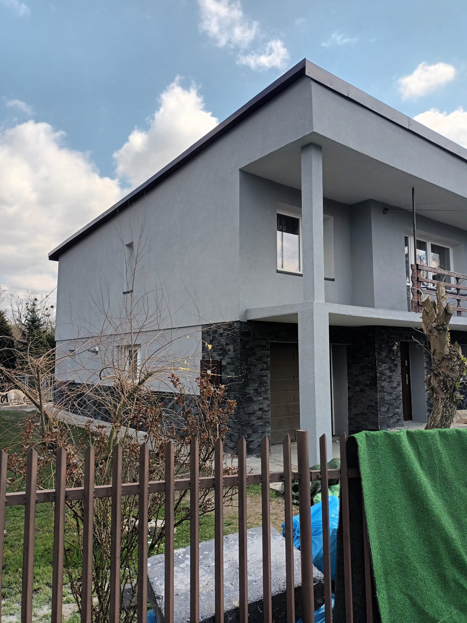 Nowoczesny, szary dom z balkonem i elewacją wykończoną kamieniem, widziany zza metalowego ogrodzenia z zielonym materiałem i niebieską folią w tle.