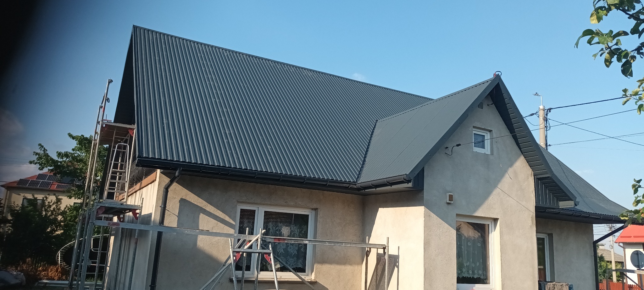 Nowy, ciemnoszary dach z blachy trapezowej na jasnym domu jednorodzinnym, widoczne rusztowanie przy ścianie bocznej, błękitne niebo w tle.