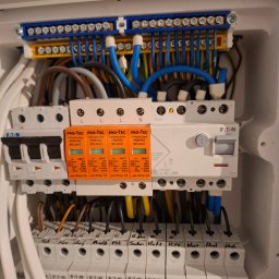 Rafał Widomski - Rozdzielnica elektryczna z zabezpieczeniami przepięciowymi Pro-Tec i wyłącznikami Eaton, widoczne przewody w kolorach niebieskim, żółtym i brązowym.