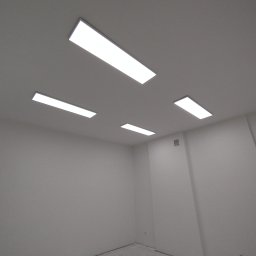 Rafał Widomski - Białe, prostokątne panele LED w suficie, oświetlające puste, białe pomieszczenie. Widok pod kątem, ukazujący rozmieszczenie lamp i minimalistyczny design wnętrza.