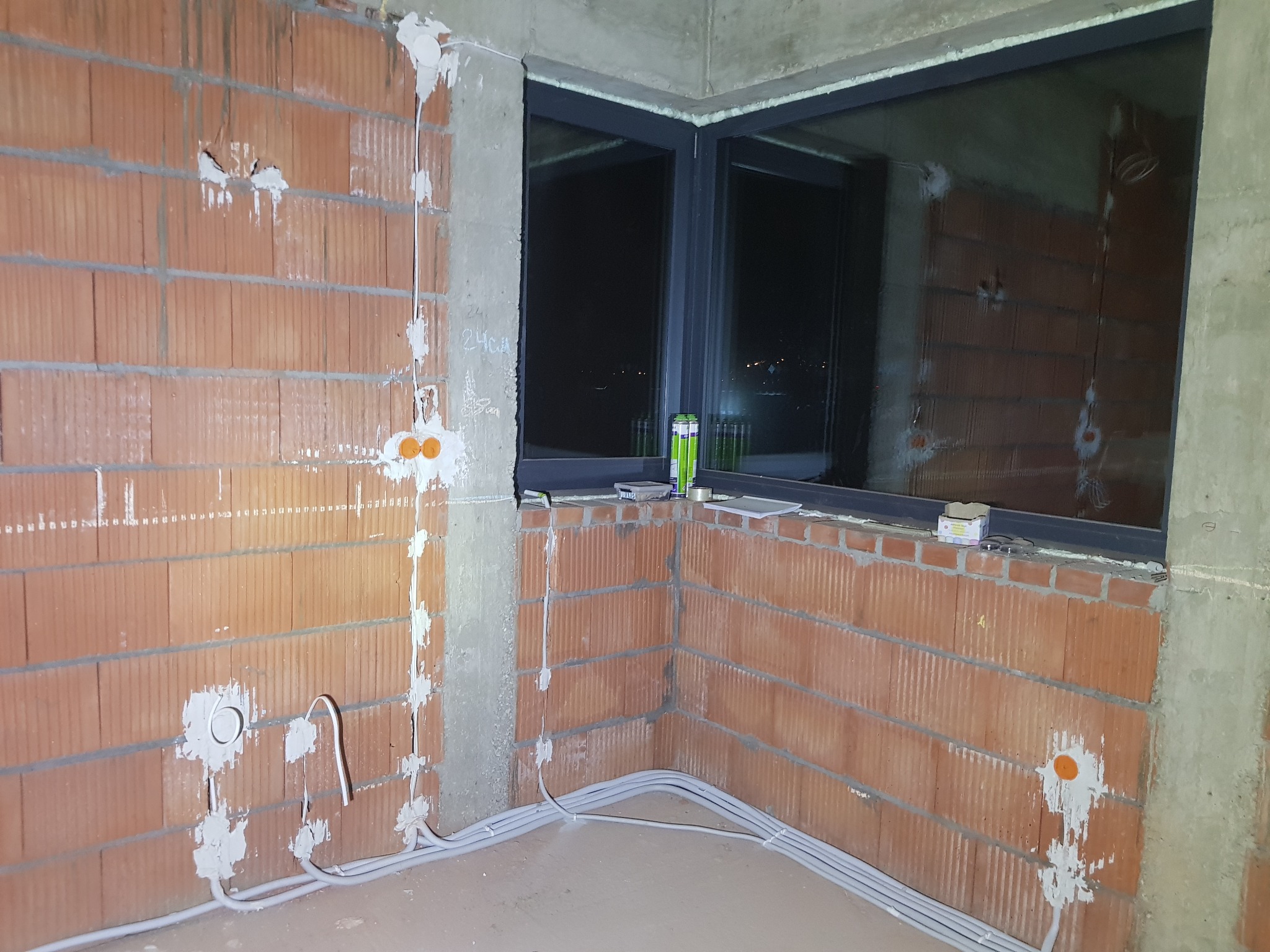 Surowy mur z instalacją elektryczną w trakcie montażu. Widoczne kable w peszlach, puszki pod gniazdka i okno z widokiem na nocne miasto.