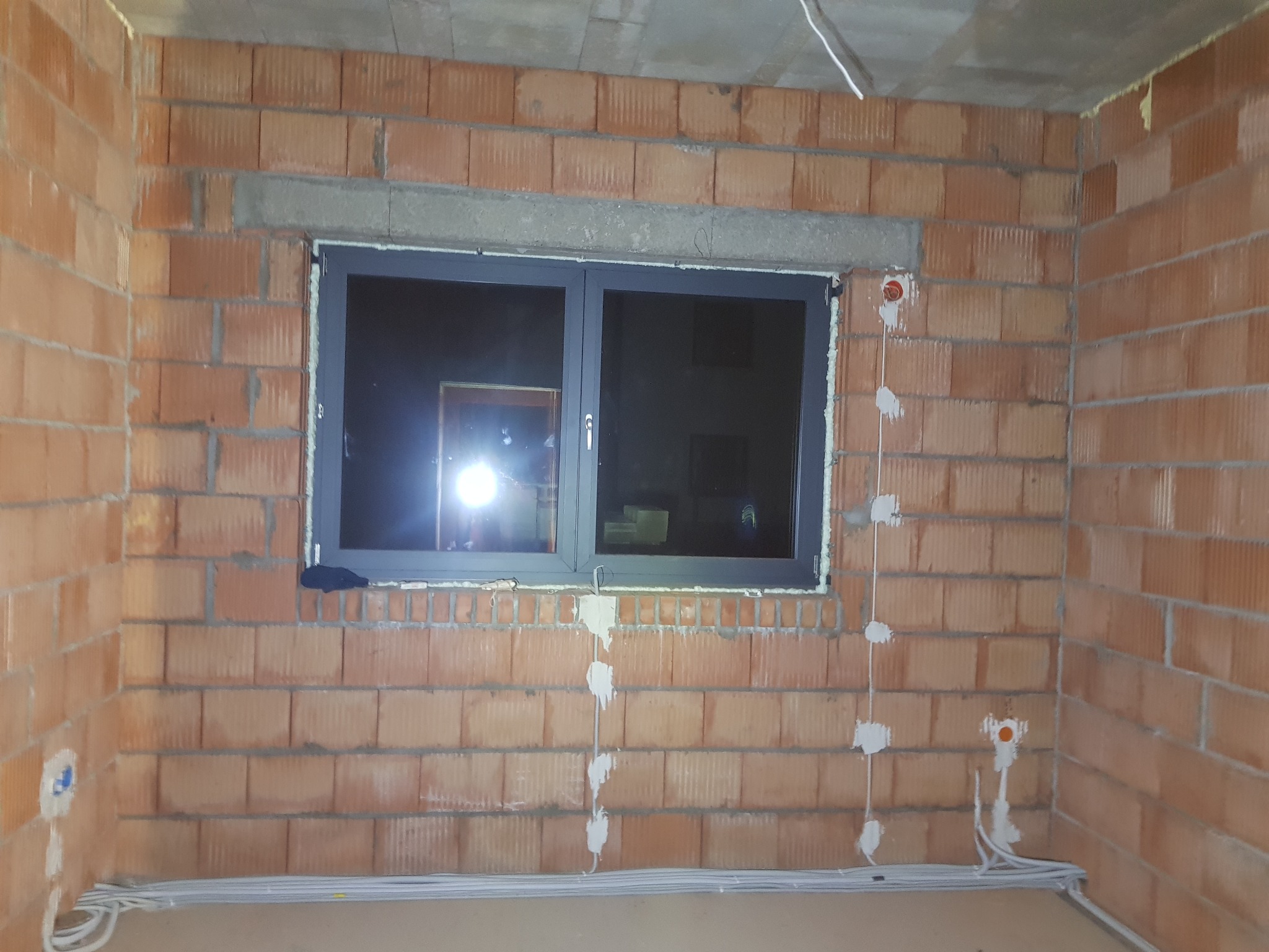 Wewnętrzna ściana z cegły z oknem i przygotowanymi punktami elektrycznymi, kable ułożone wzdłuż ściany. Widoczne puszki instalacyjne.