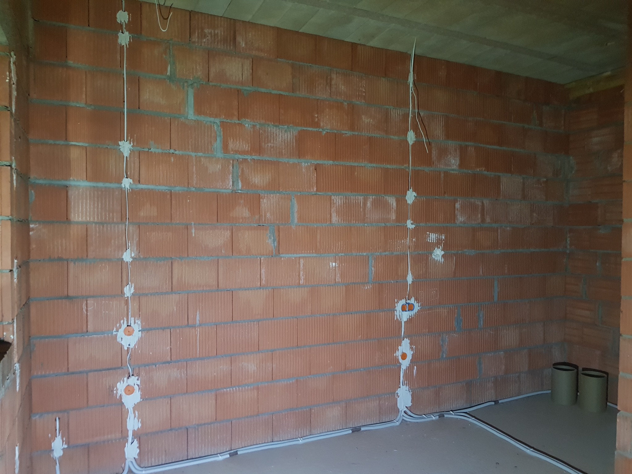Instalacja elektryczna w trakcie montażu, widoczne kable prowadzone po ścianie z cegły, puszki instalacyjne zamontowane w ścianie, surowy stan.