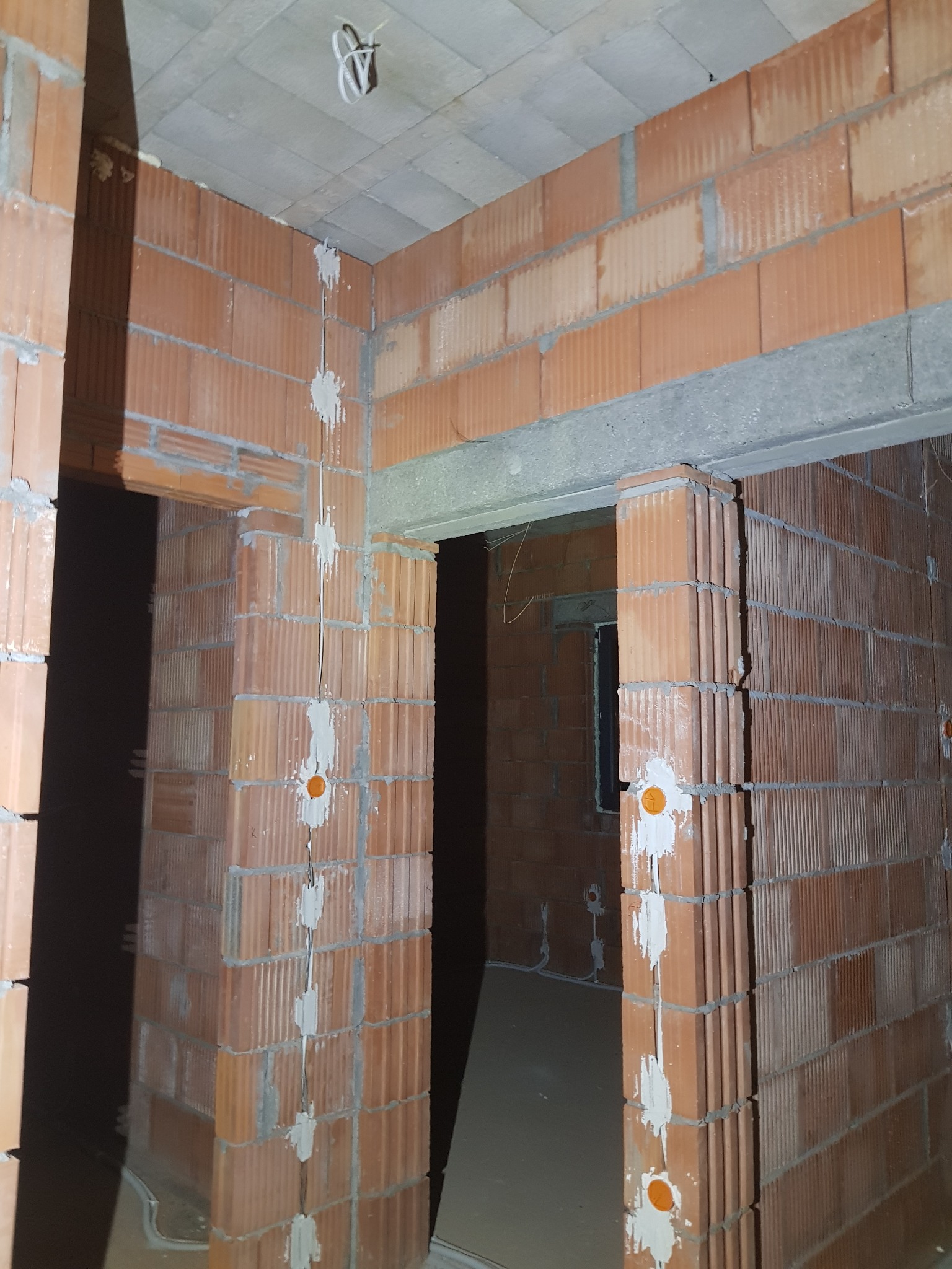 Murowane ściany z widocznymi punktami instalacji elektrycznej wewnątrz budynku w stanie surowym, przewody zabezpieczone gipsem, widoczne kable pod sufitem.